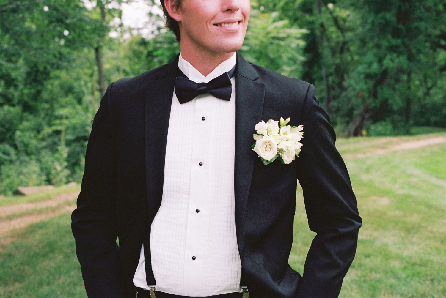 Smiling groom in black tuxedo with white boutonniere and bow tie standing outdoors.