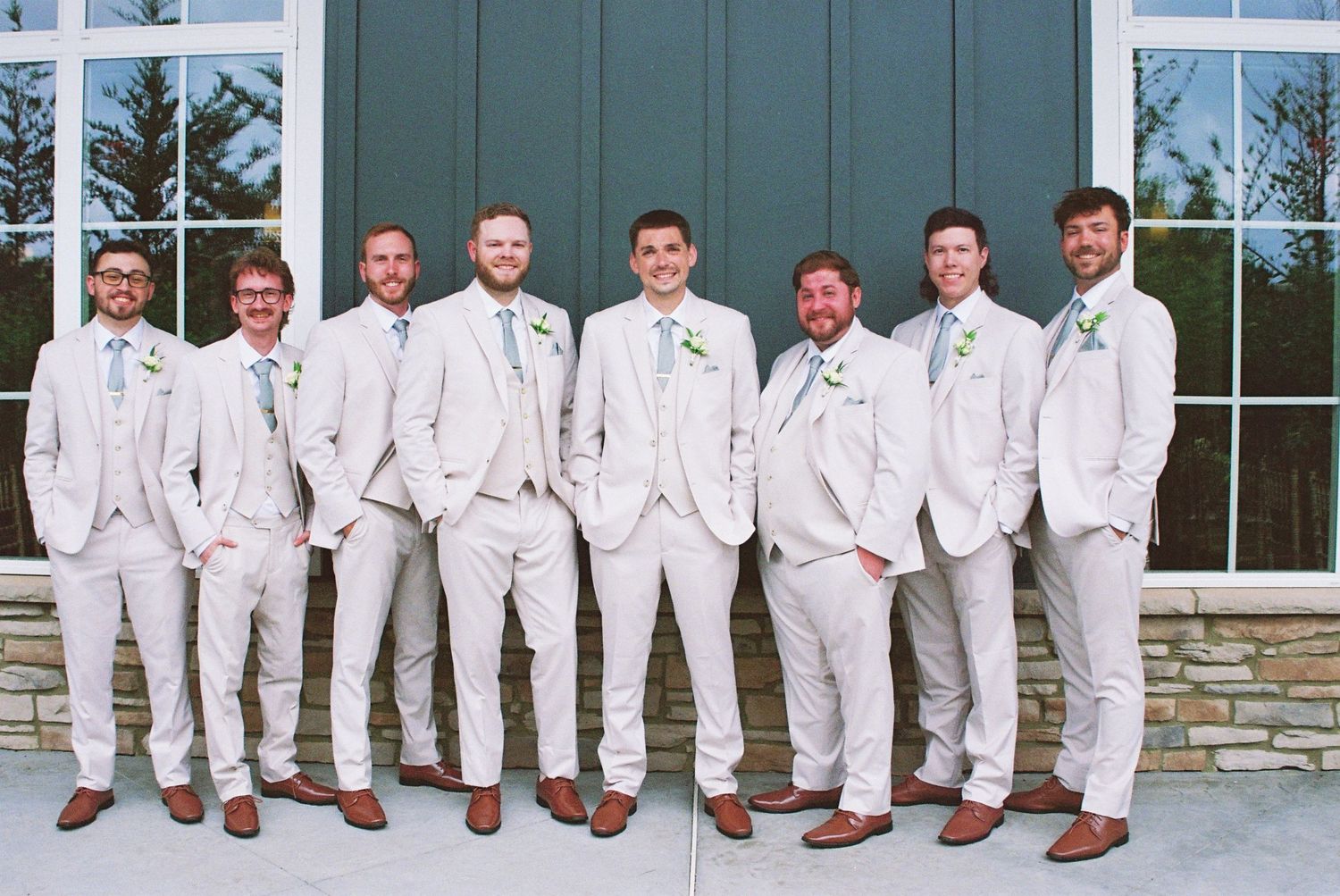 Groom and eight groomsmen in matching light gray suits posing in front of a dark blue barn.