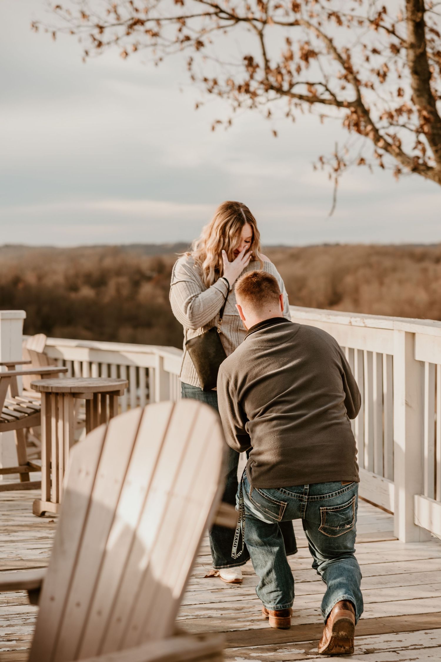 Emotional proposal scene on a wooden observation deck with fall foliage in background.