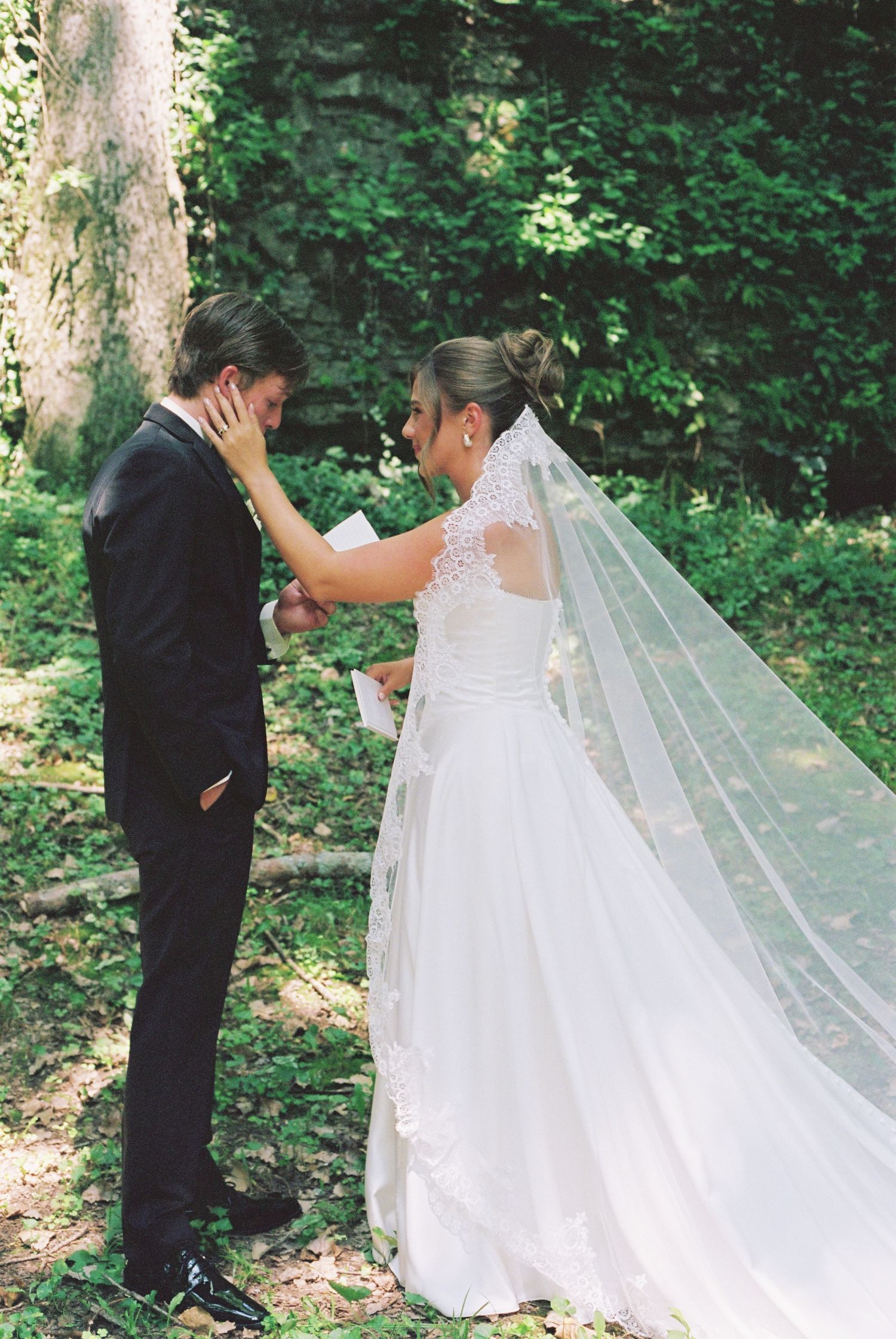 Bride in white gown and veil touches groom's face as they share an emotional first look moment in a lush forest.