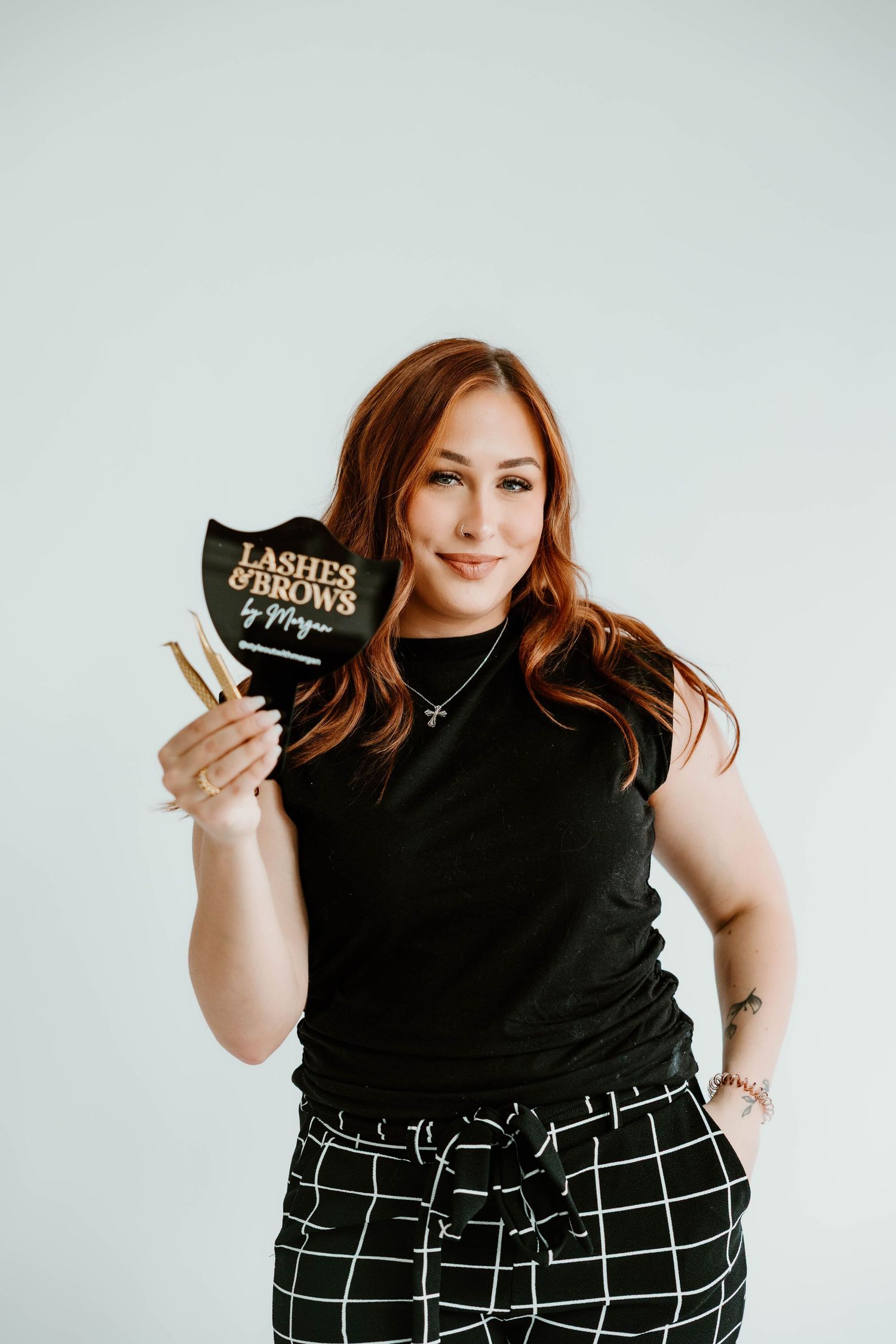 Plus size model in black top and checkered pants showing makeup products against white background.