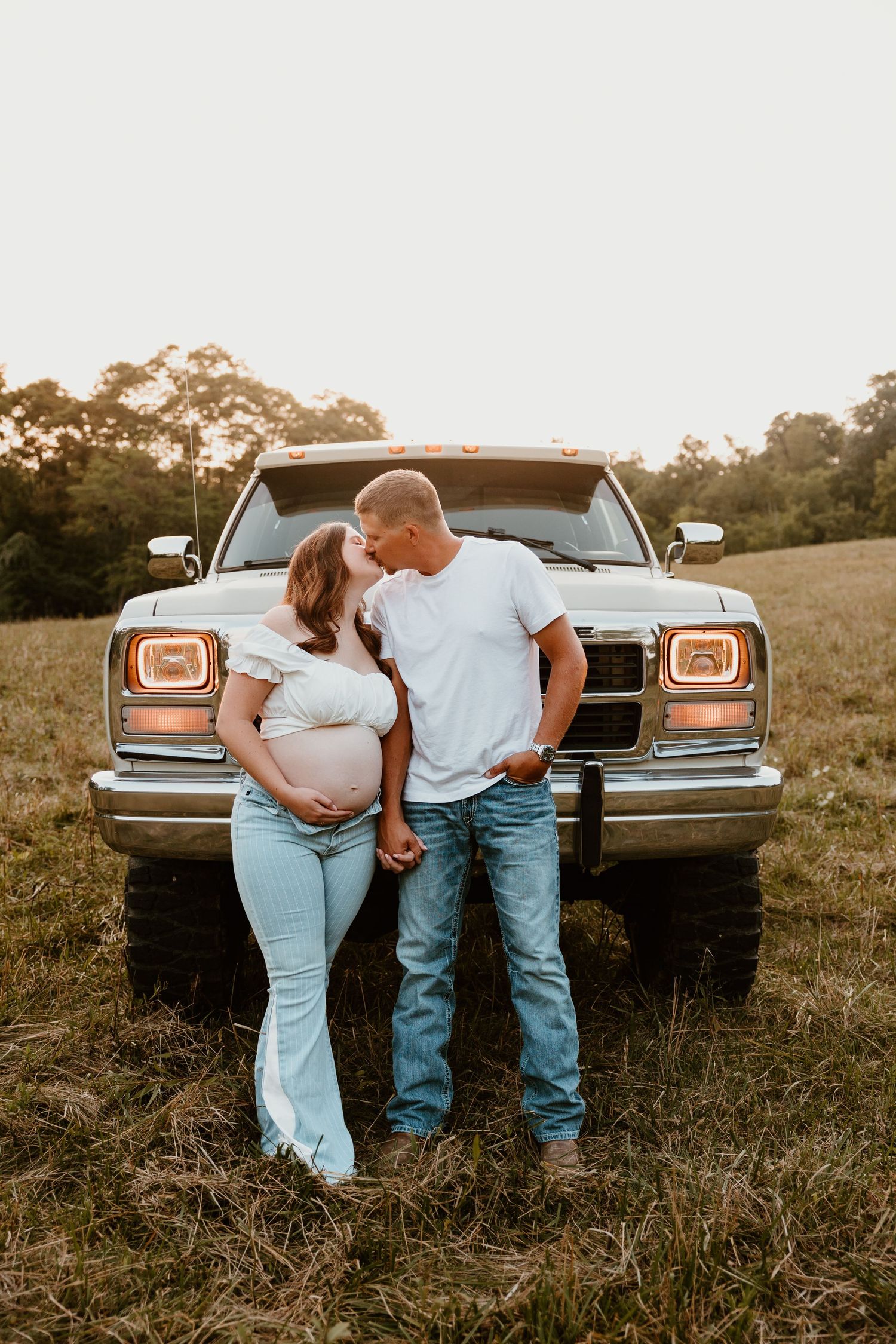 A couple poses together in front of a classic car in a rustic field at sunset.