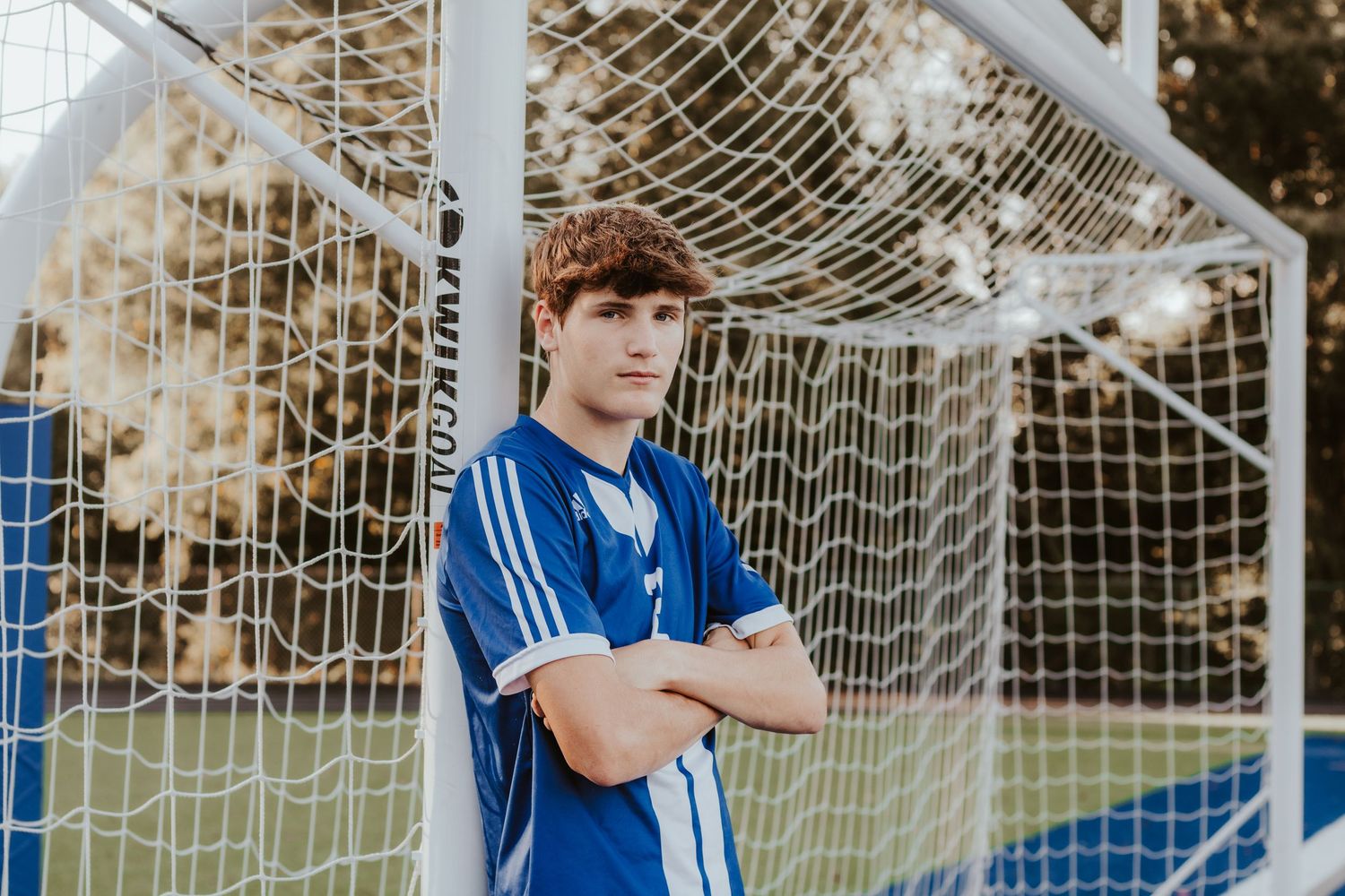 Soccer player posing by goal net in blue uniform on school field.