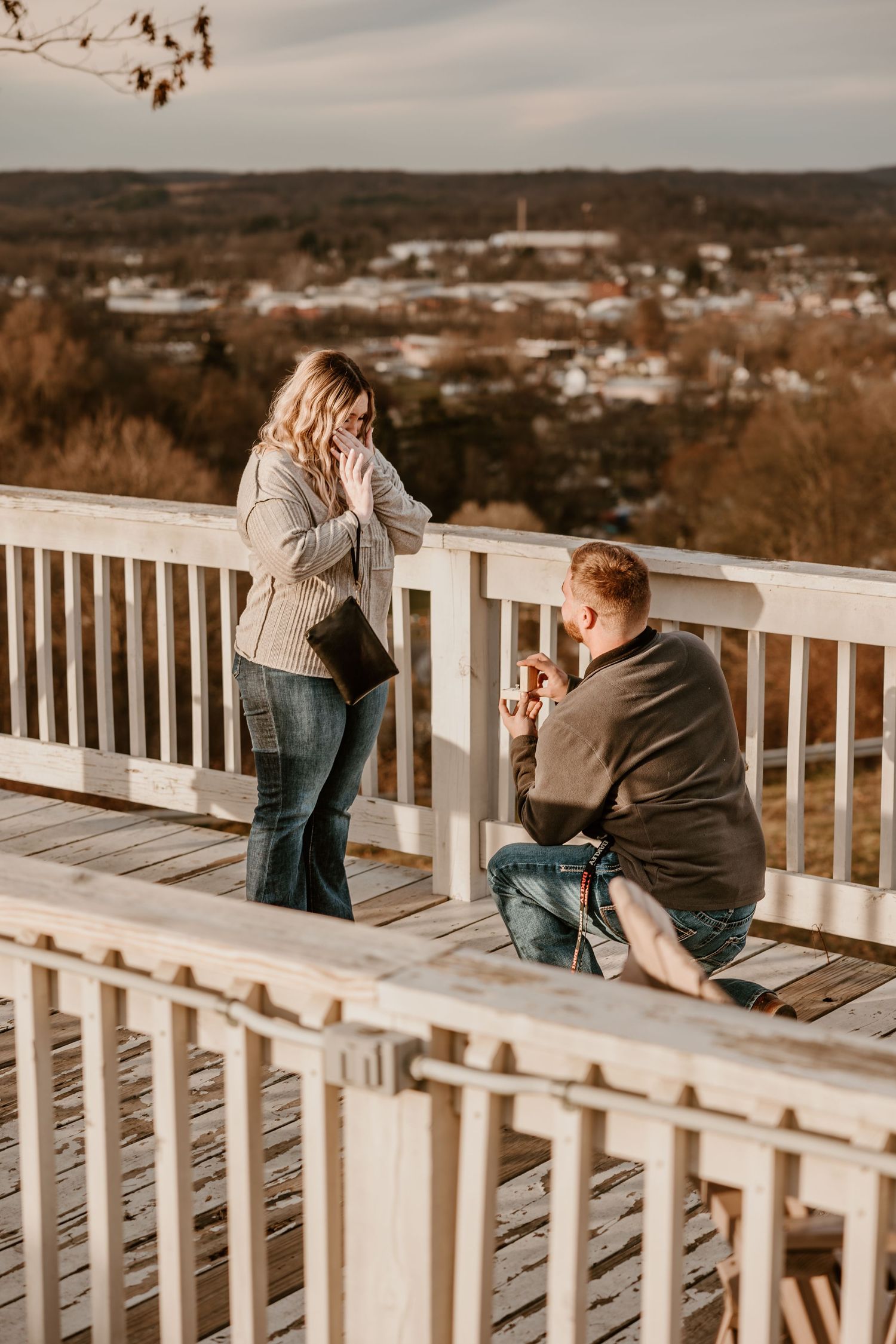 Surprise proposal moment captured on a scenic wooden deck overlooking a town.