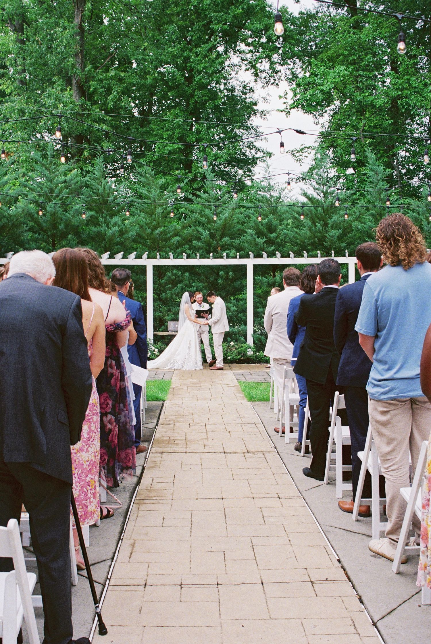Outdoor garden wedding ceremony with guests standing as bride and groom exchange vows at altar.