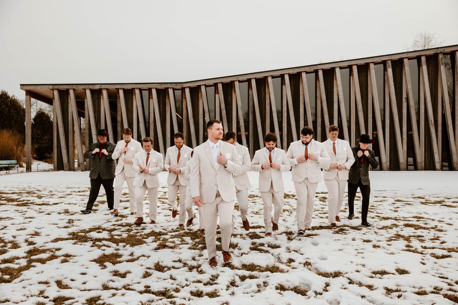 Wedding party lined up against wooden fence in winter snow scene.