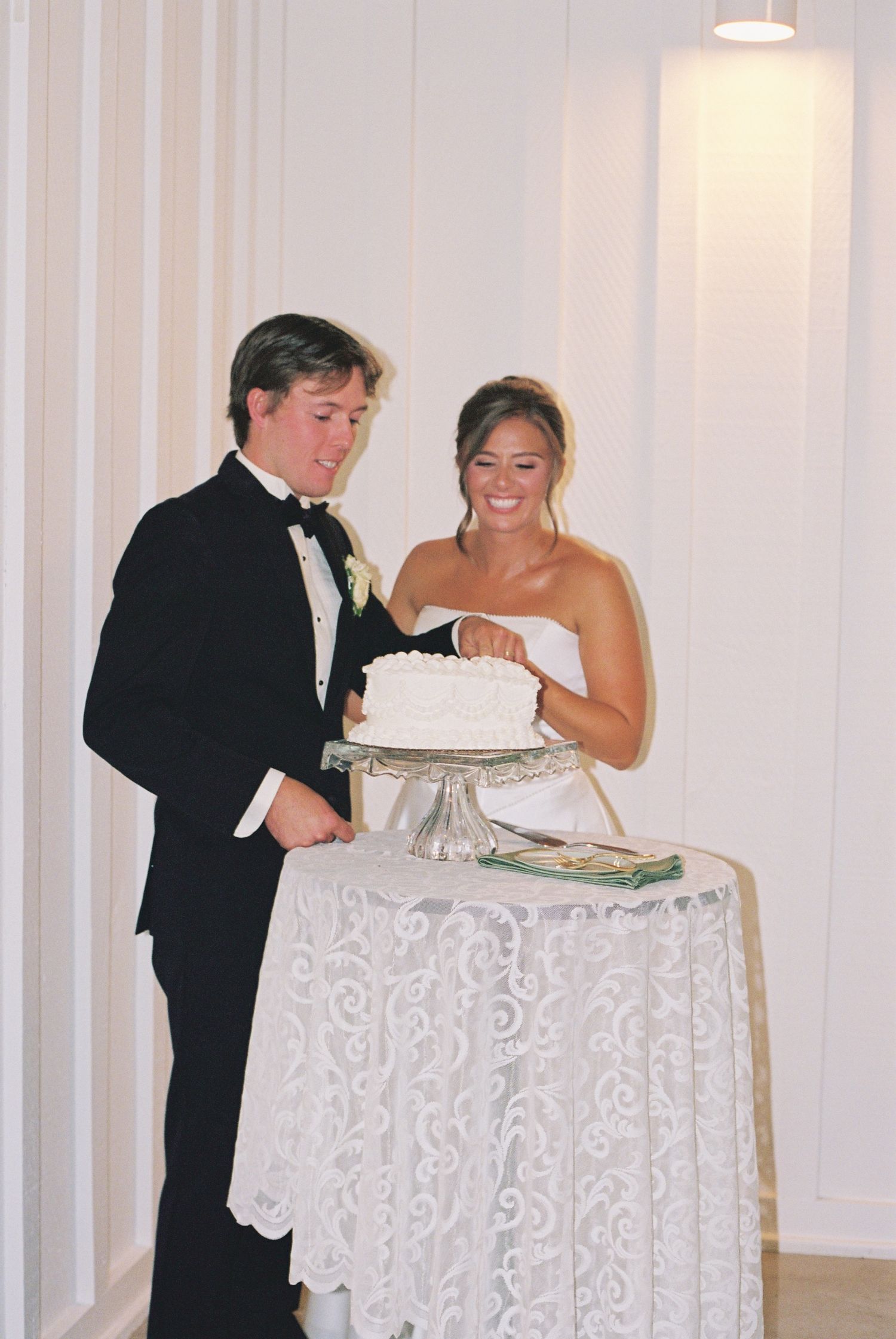 Bride and groom cutting white wedding cake on lace-covered table at elegant indoor wedding reception.