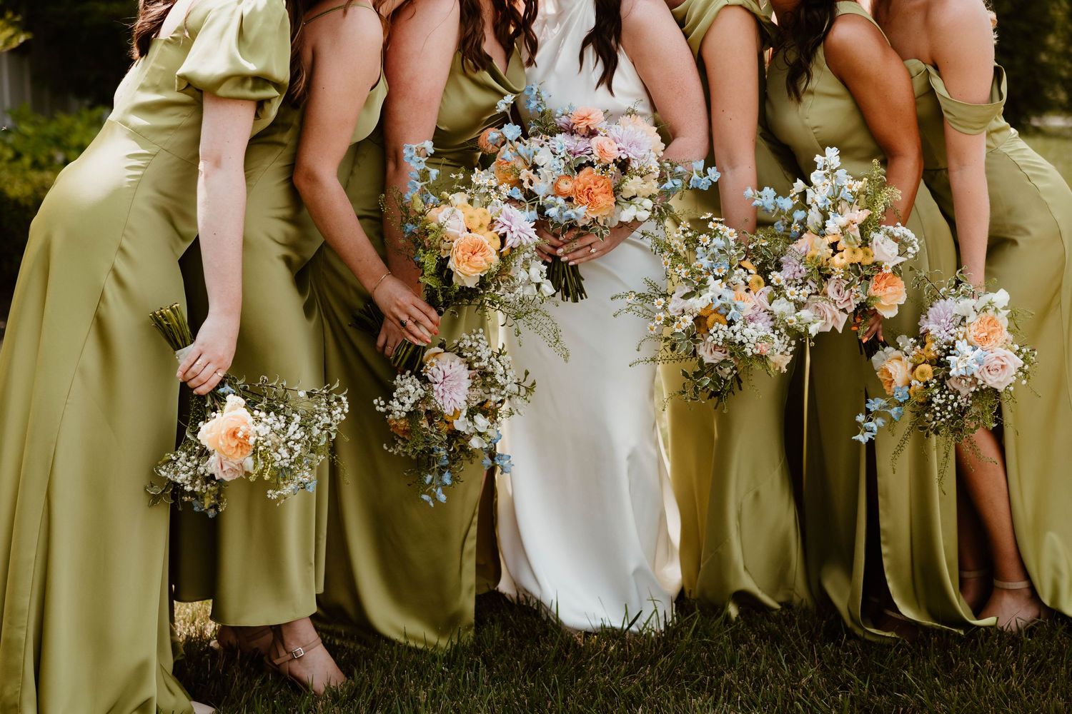 Bride in white gown with bridesmaids in olive green satin dresses holding colorful floral bouquets outdoors.