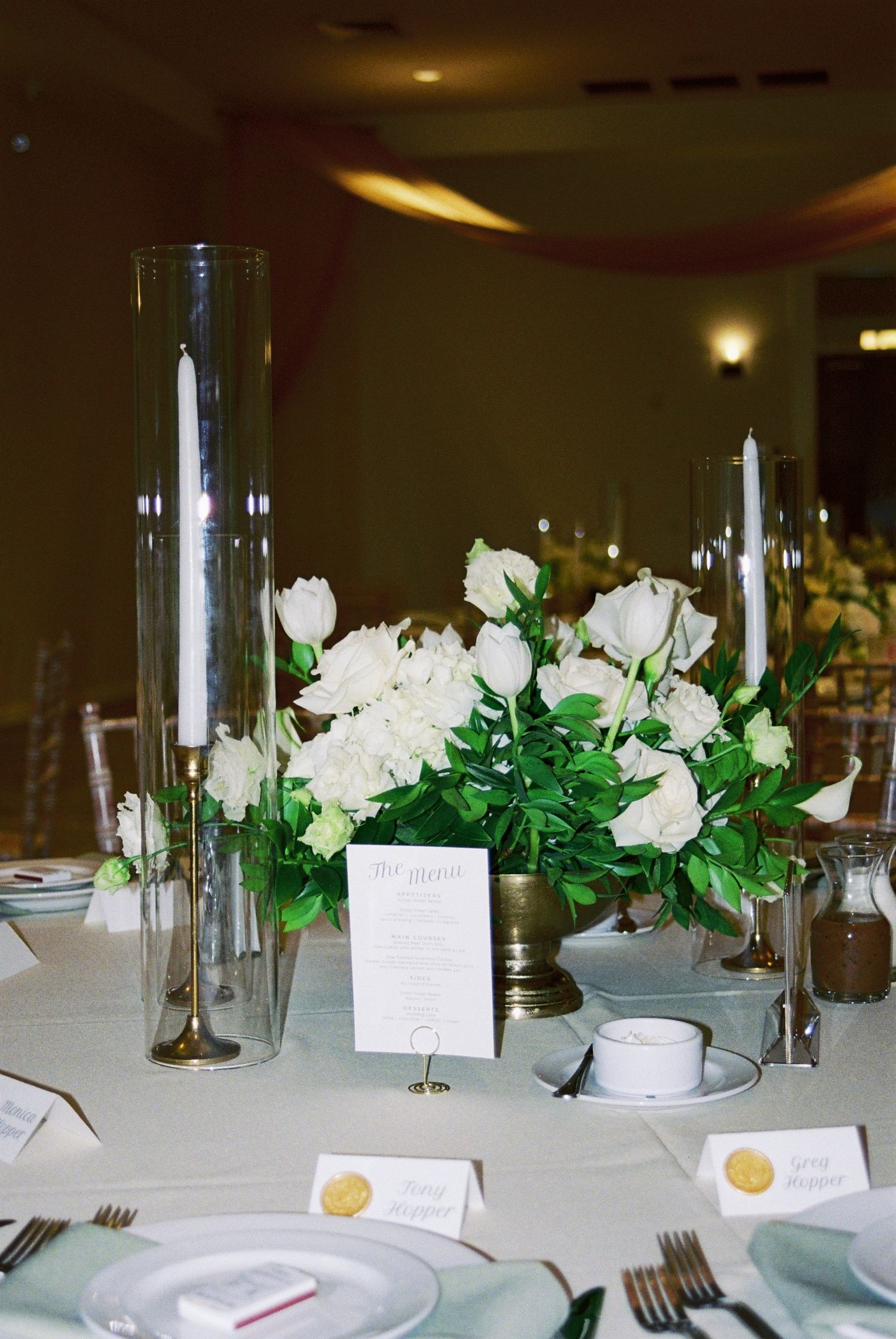 Wedding reception table centerpiece with white roses, greenery, tall glass candle holders, and menu card.