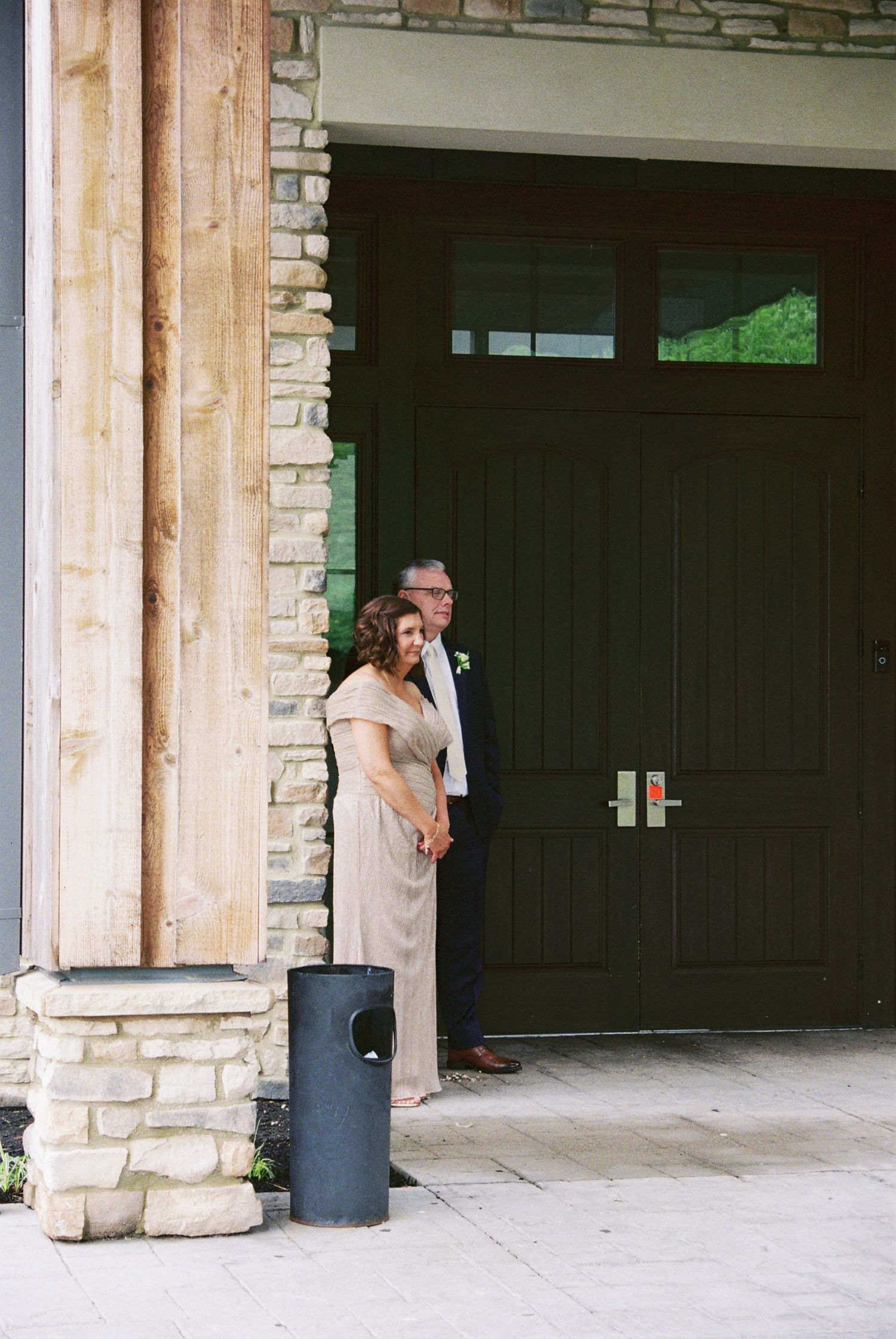 Couple in formal wedding attire standing by a dark green door of a stone and wood building.