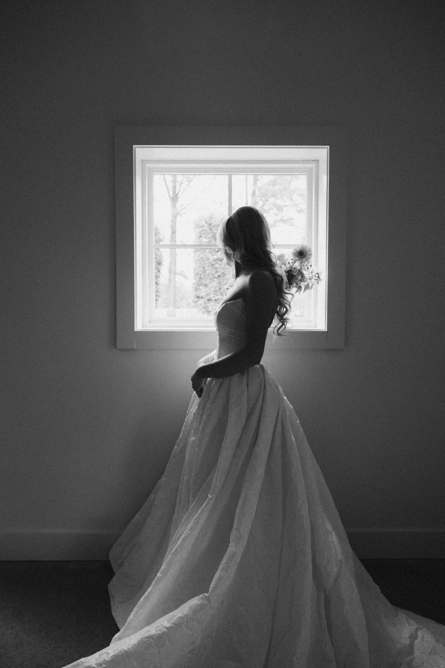 Black and white photo of a bride in a flowing wedding gown holding bouquet, standing by a bright window.