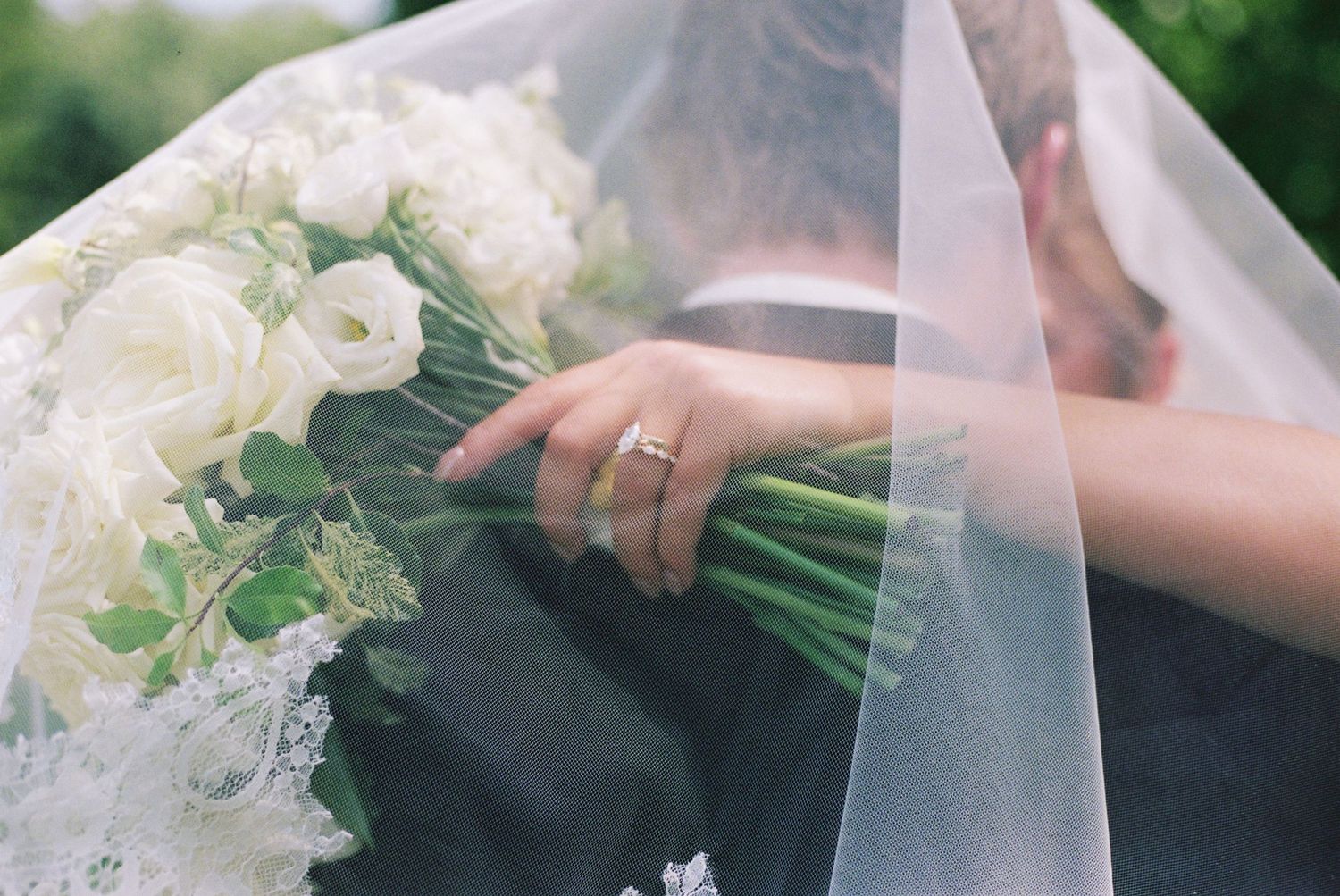 Bride holding white rose wedding bouquet with gold ring visible under sheer veil.