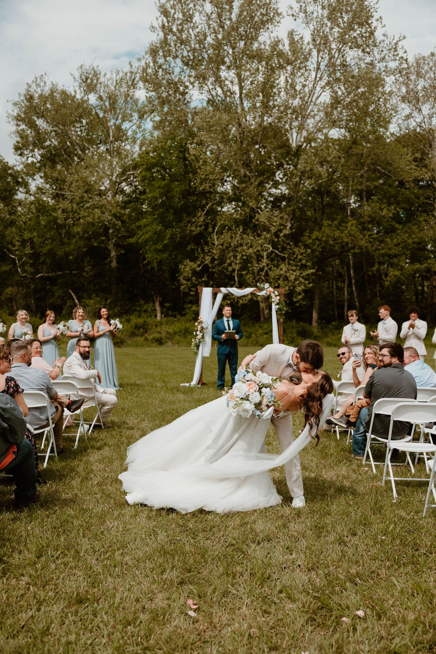 Groom dips bride for a kiss during outdoor wedding ceremony on green lawn with wedding party and guests.