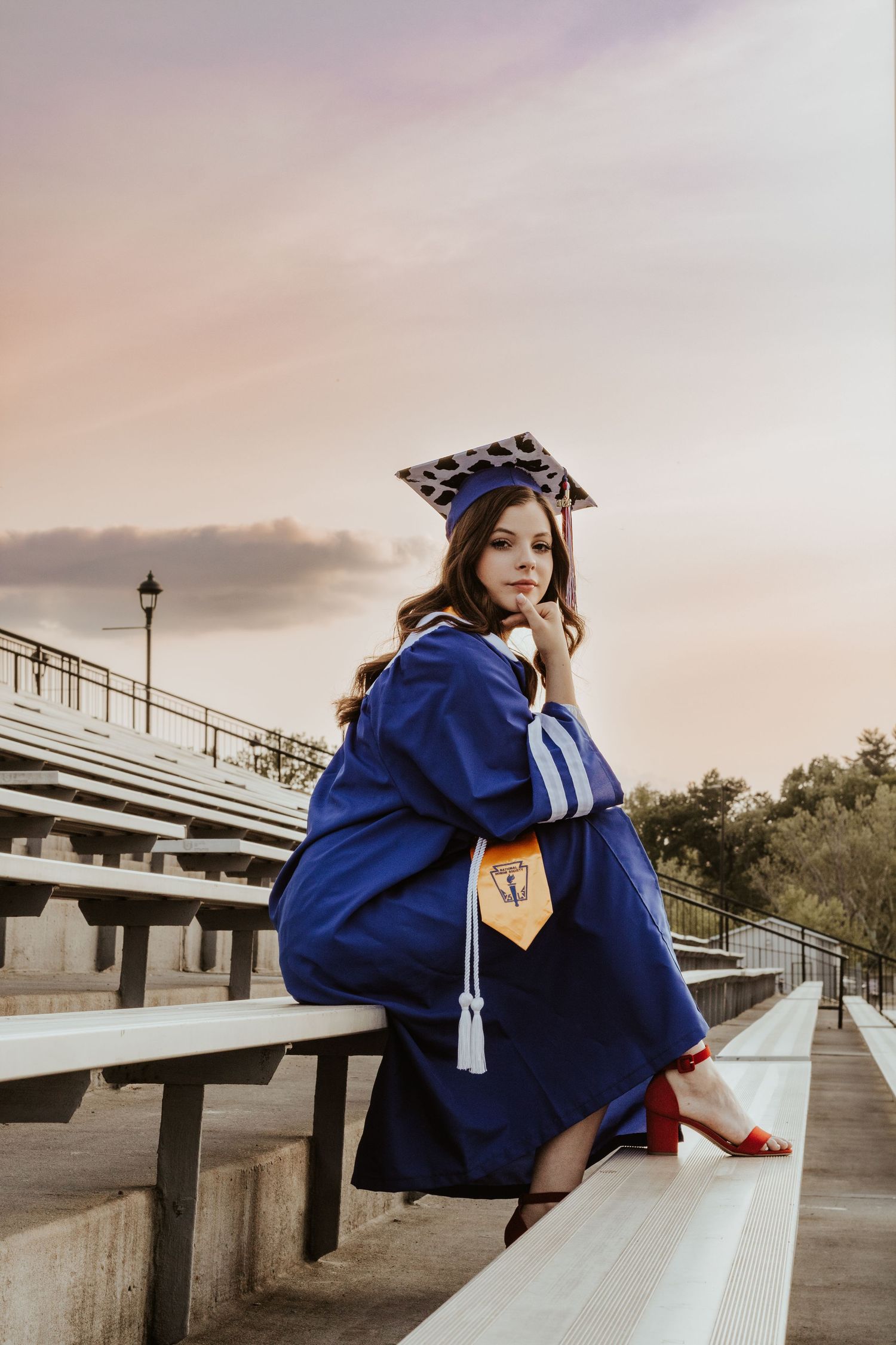 Graduate in blue cap and gown sitting on concrete bleachers at sunset.