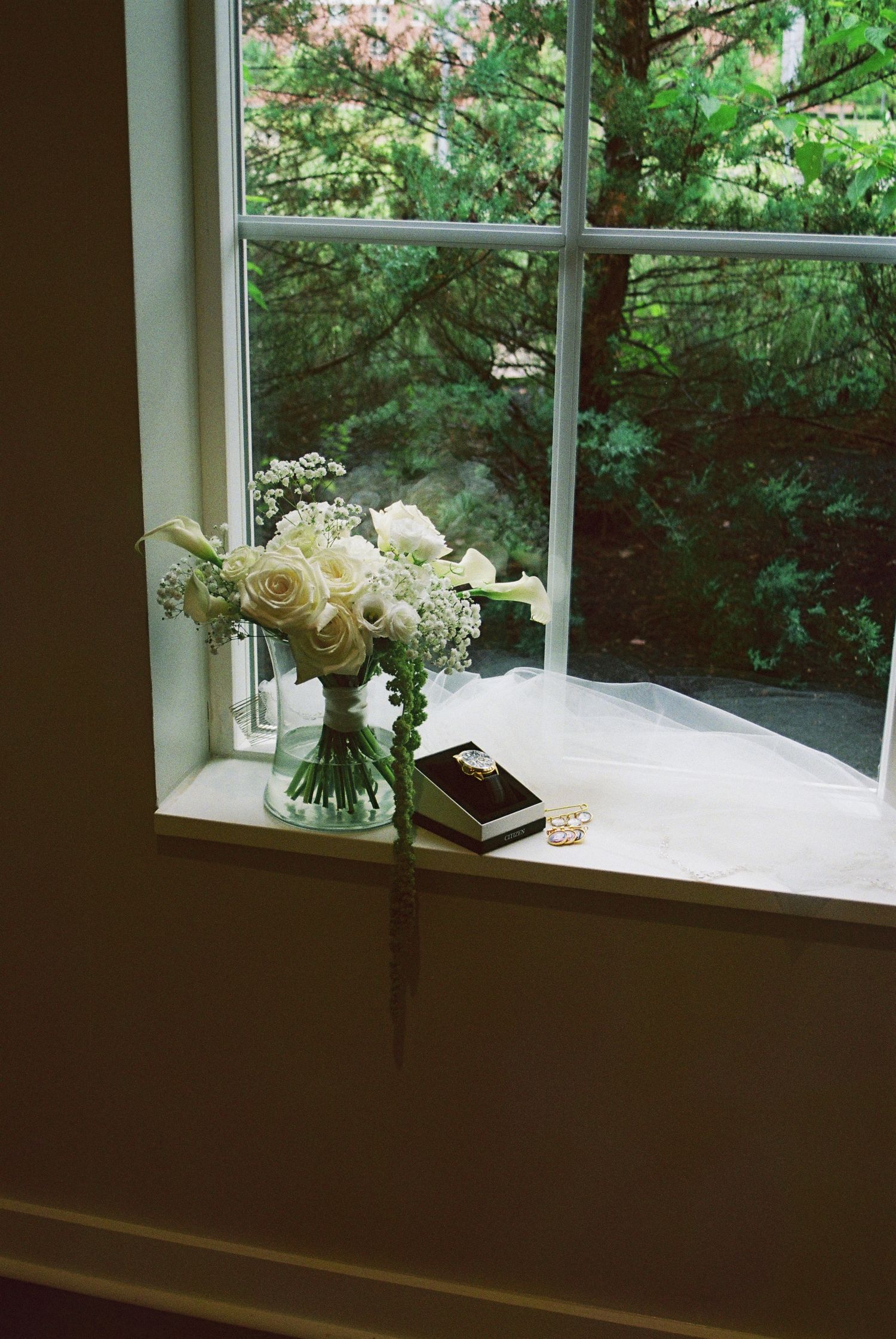 White rose and calla lily bridal bouquet displayed on windowsill with wedding rings and greenery backdrop.