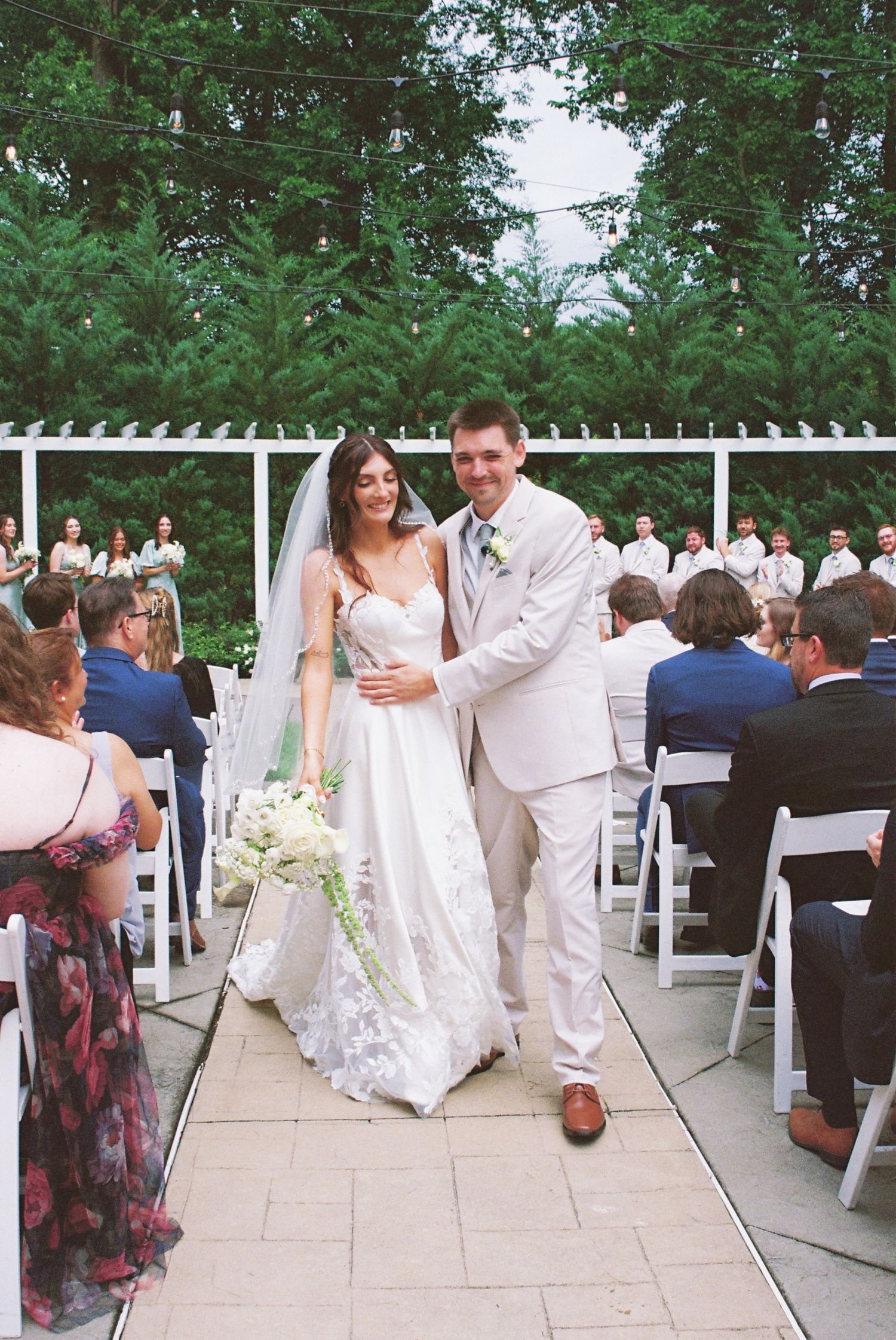 Smiling newlyweds walk back down the aisle after outdoor garden wedding ceremony with guests seated.