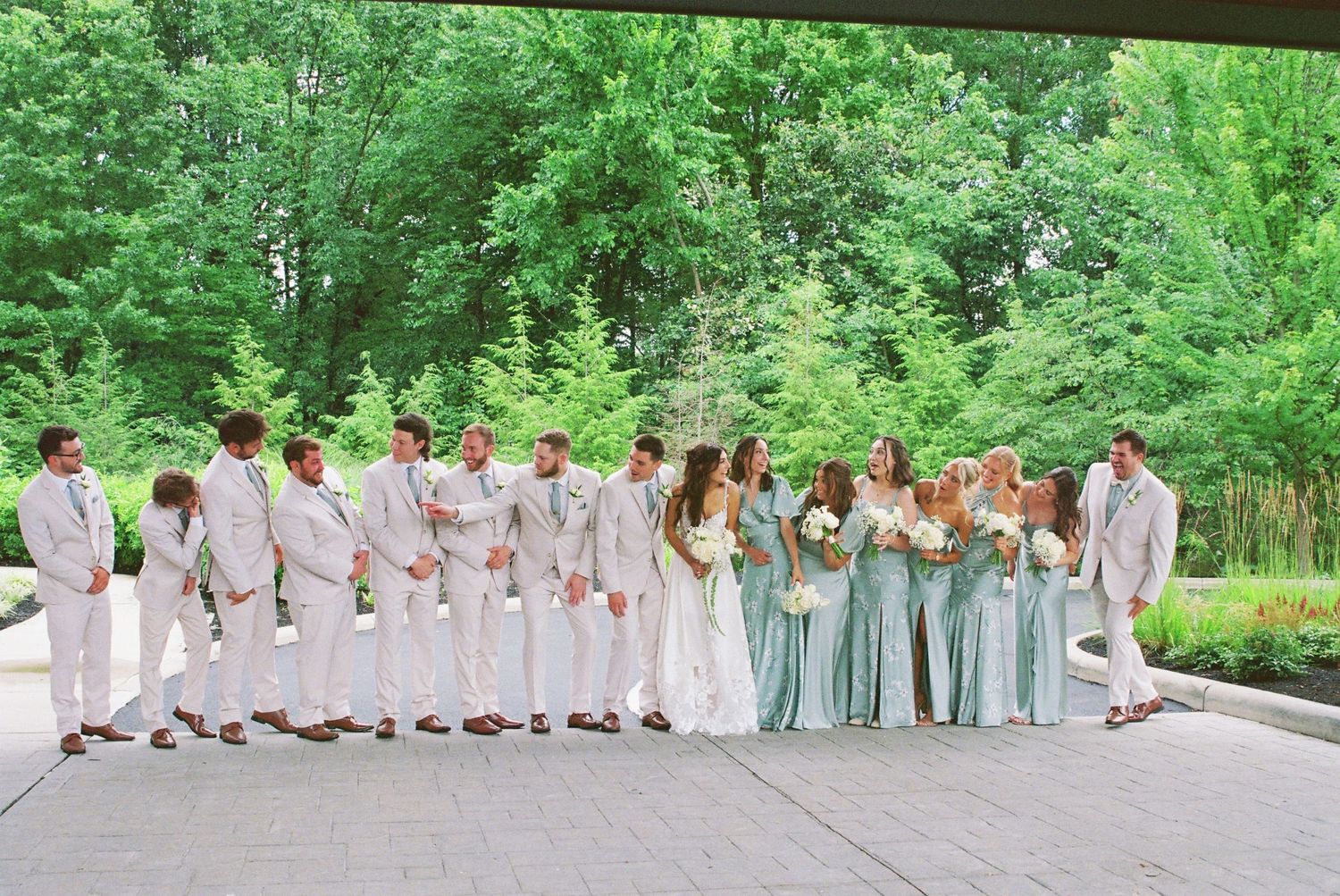 Wedding party poses outdoors in sage green bridesmaid dresses and tan suits with lush green trees in background.