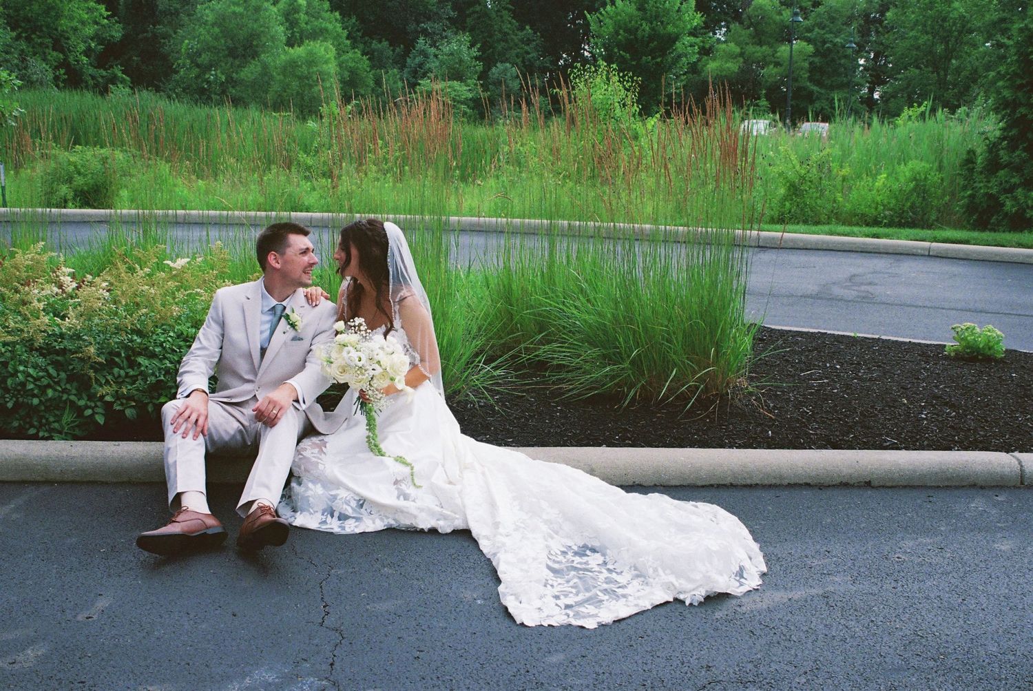 A bride and groom sit together on a curb in wedding attire, surrounded by lush green landscaping on their wedding day.