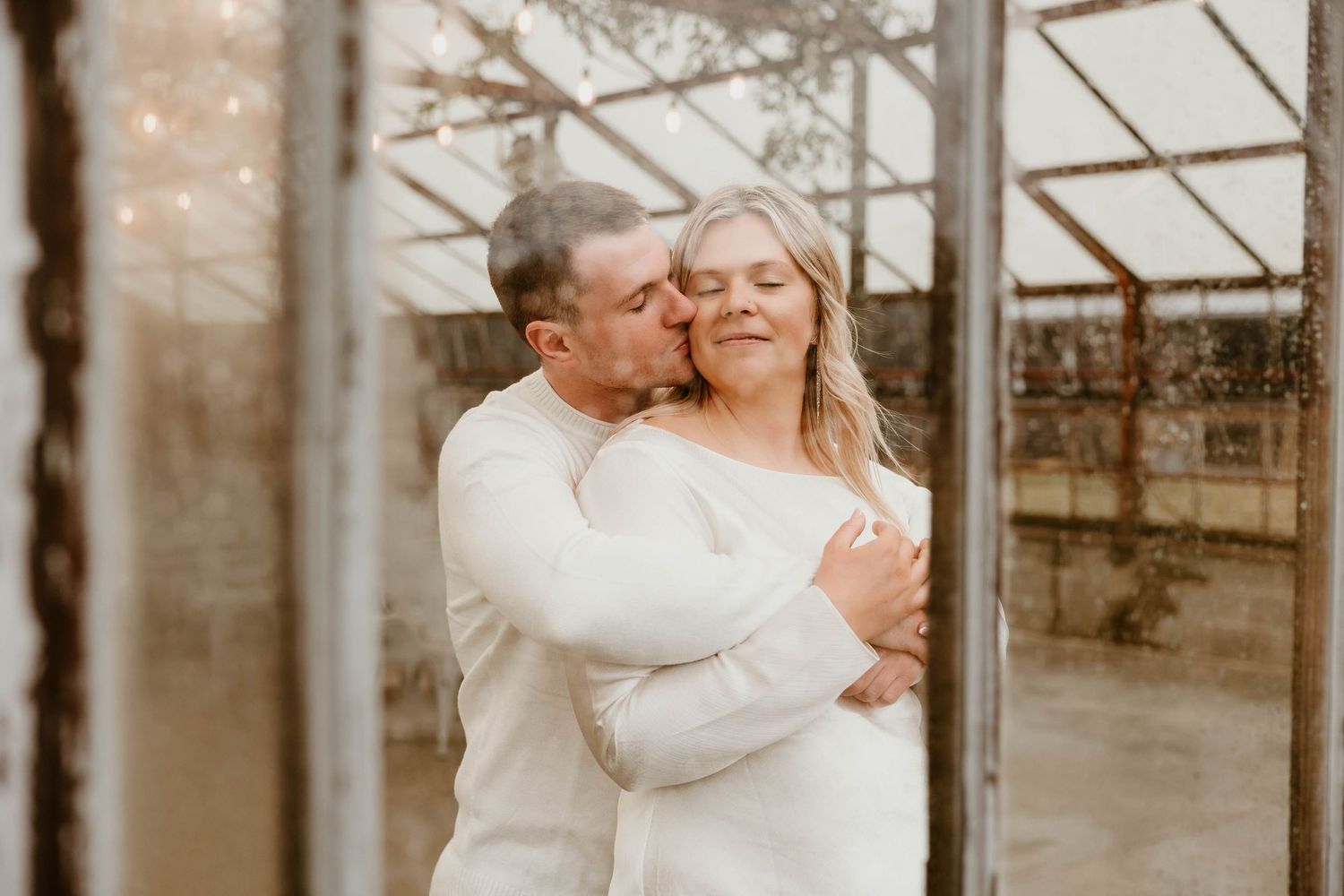 A romantic couple embraces in a greenhouse with soft lighting creating an intimate atmosphere.