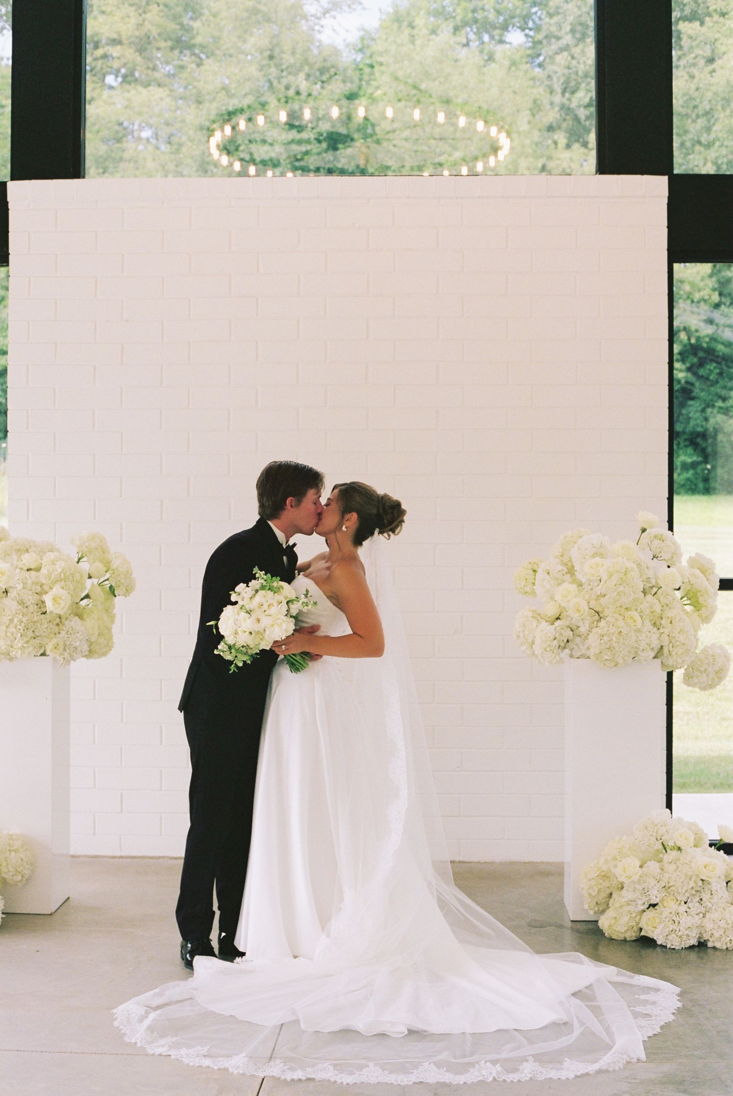 Bride and groom share first kiss at elegant white brick altar adorned with lush white hydrangea floral arrangements.
