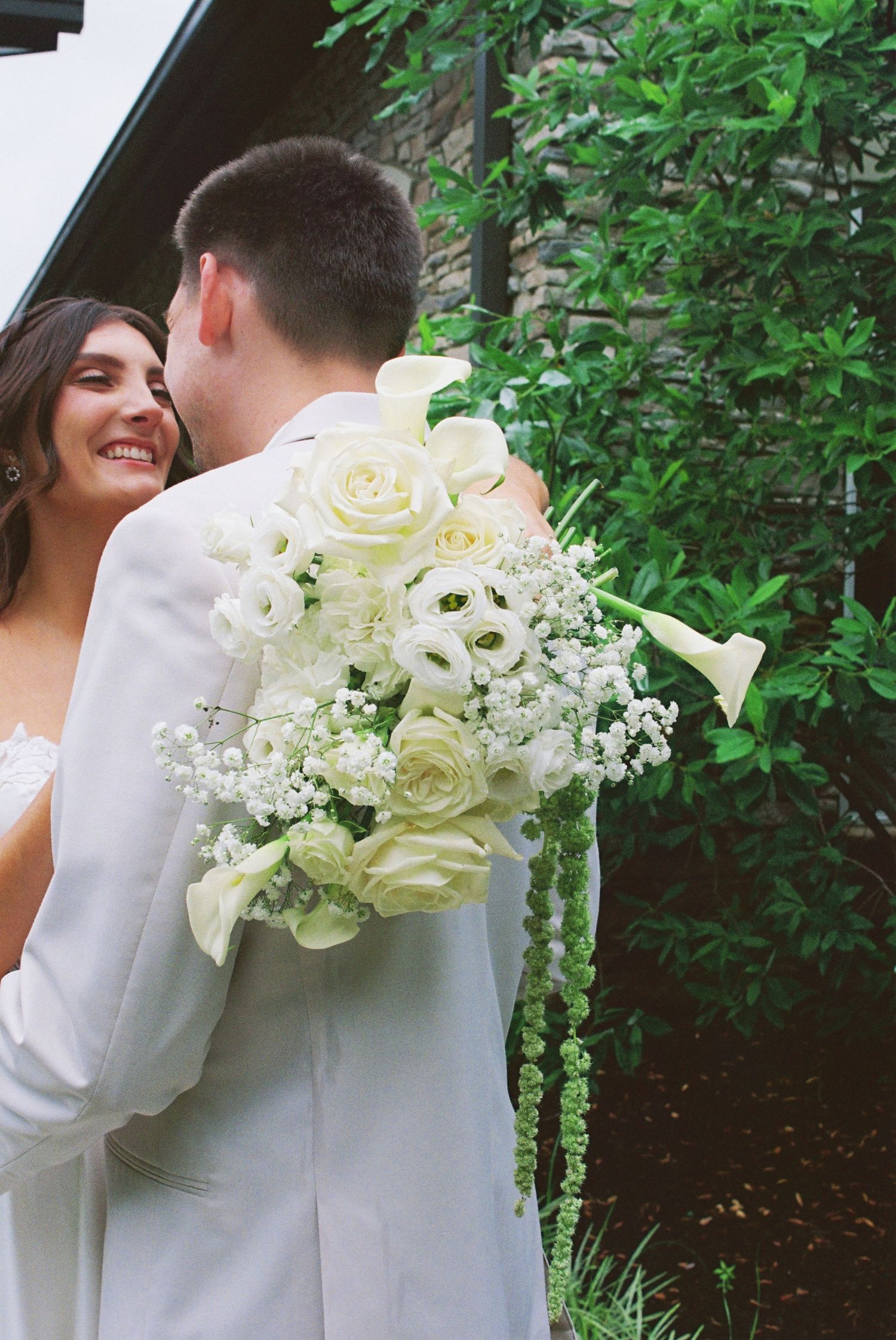 A bride smiling and embracing a groom in a white suit, holding a lush white rose and calla lily wedding bouquet outdoors.