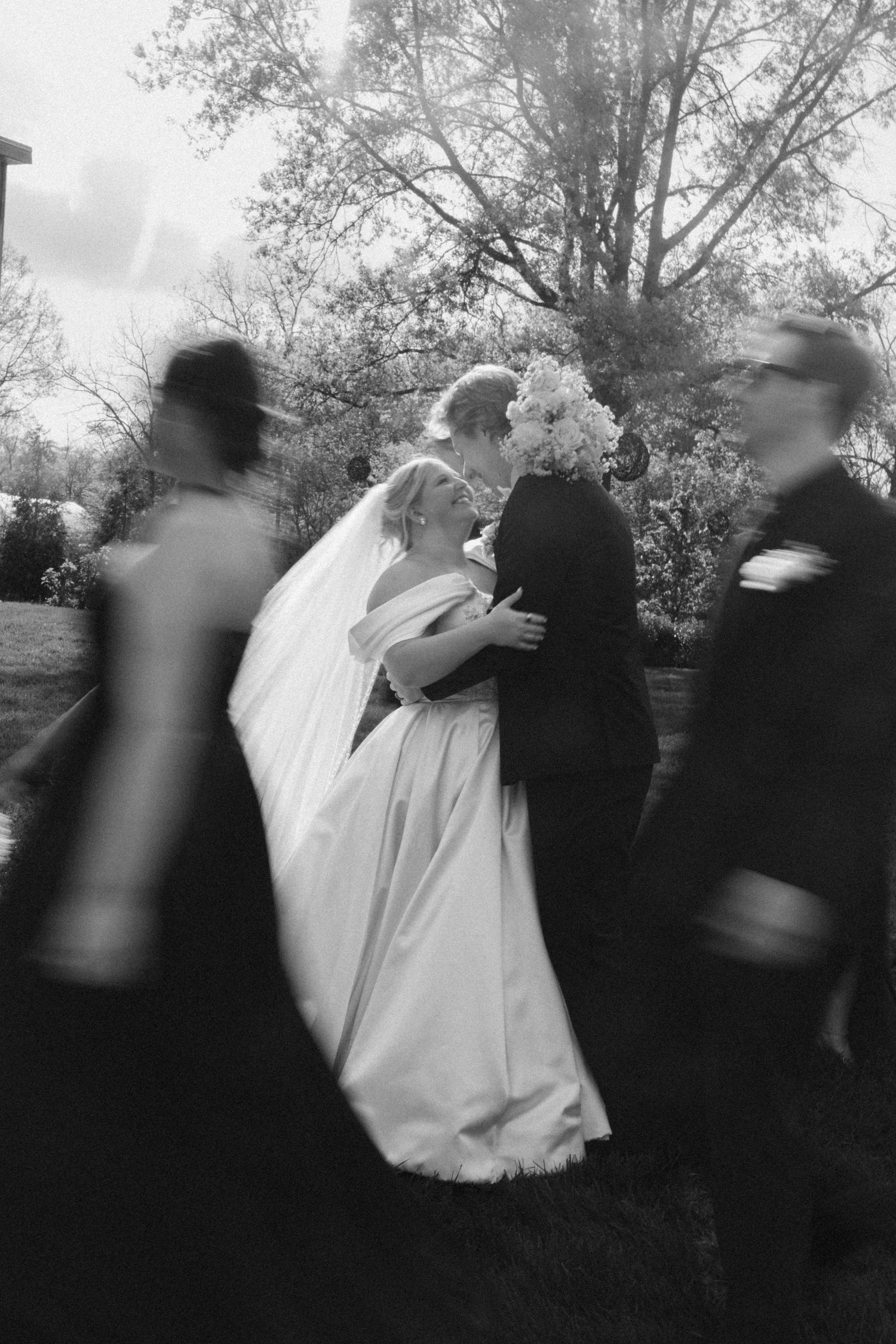 Black and white photo of a bride and groom kissing outdoors while wedding guests walk past in a blur.