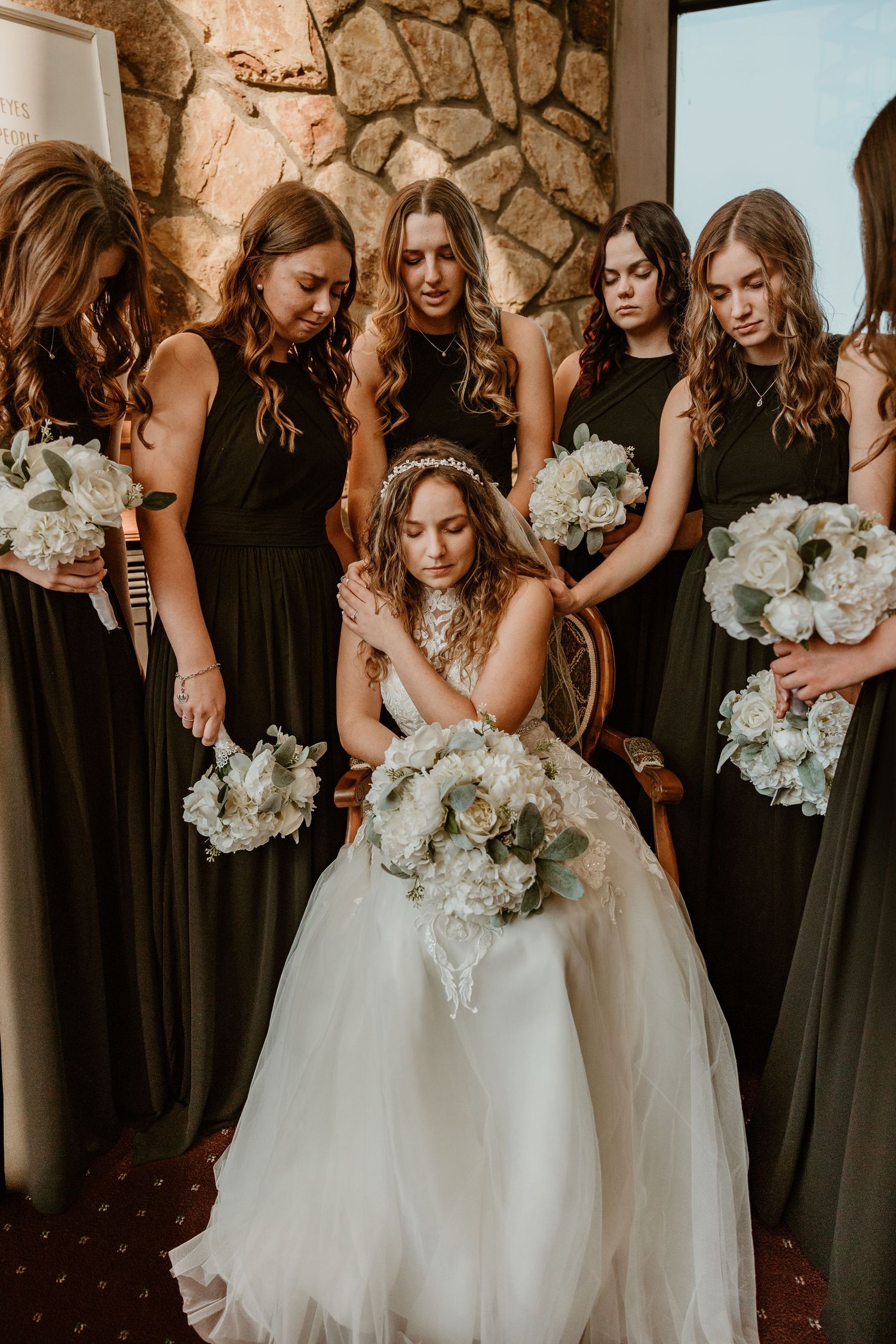 Bridesmaids in black dresses hold white floral bouquets against stone wall.