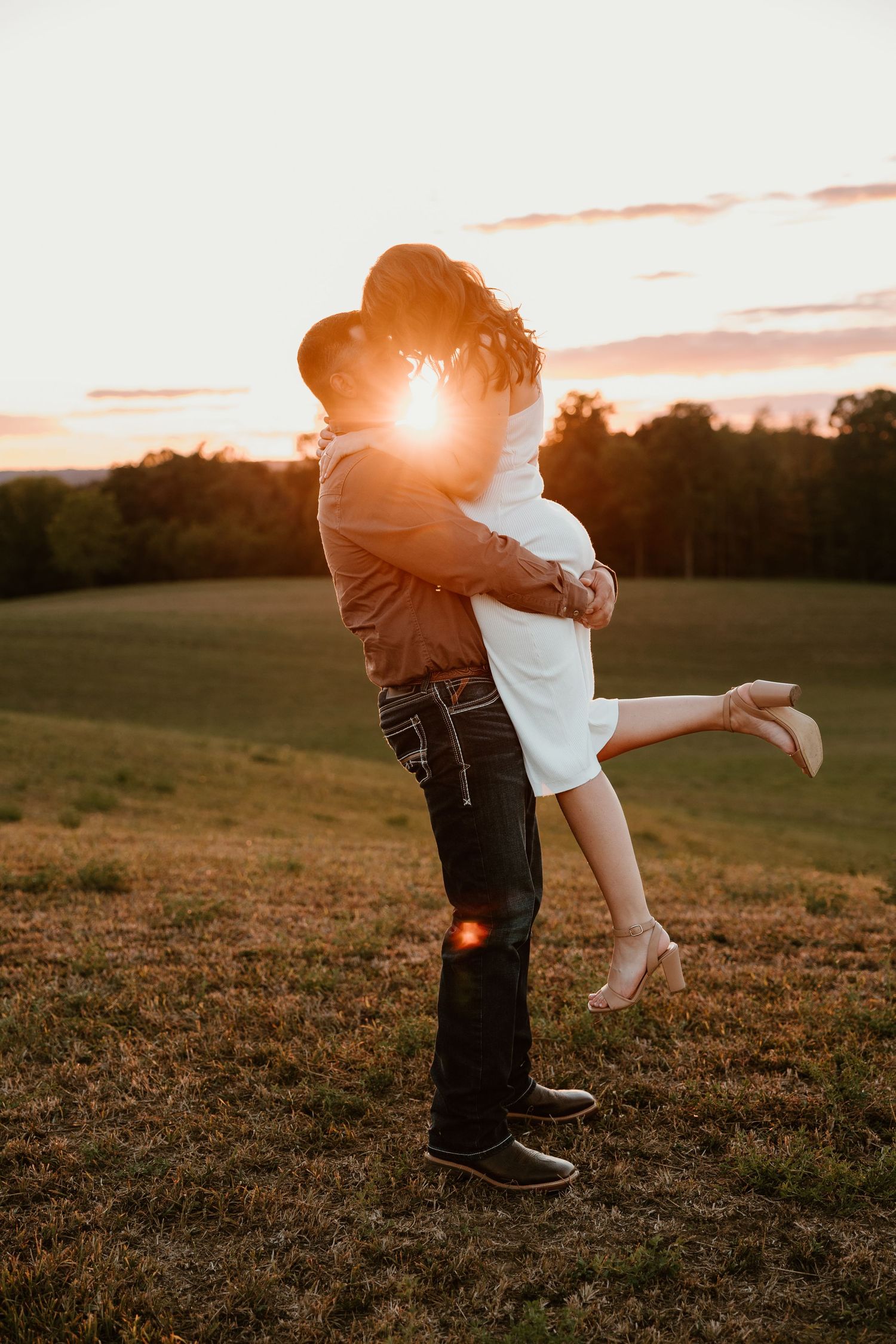 A couple embraces in a field during sunset with a golden sun flare.