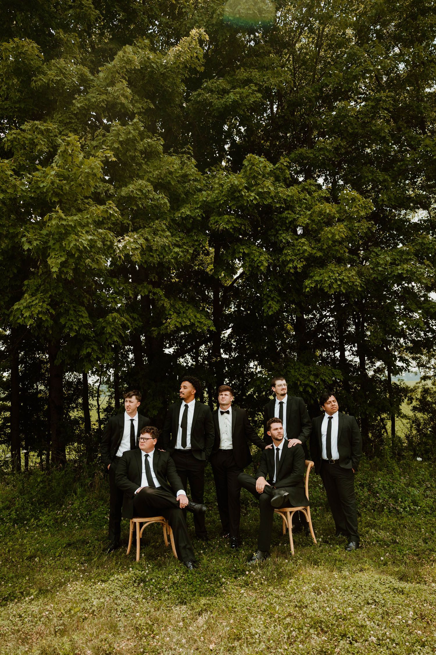Groomsmen in black suits and ties pose outdoors among lush green trees on a grassy hill.