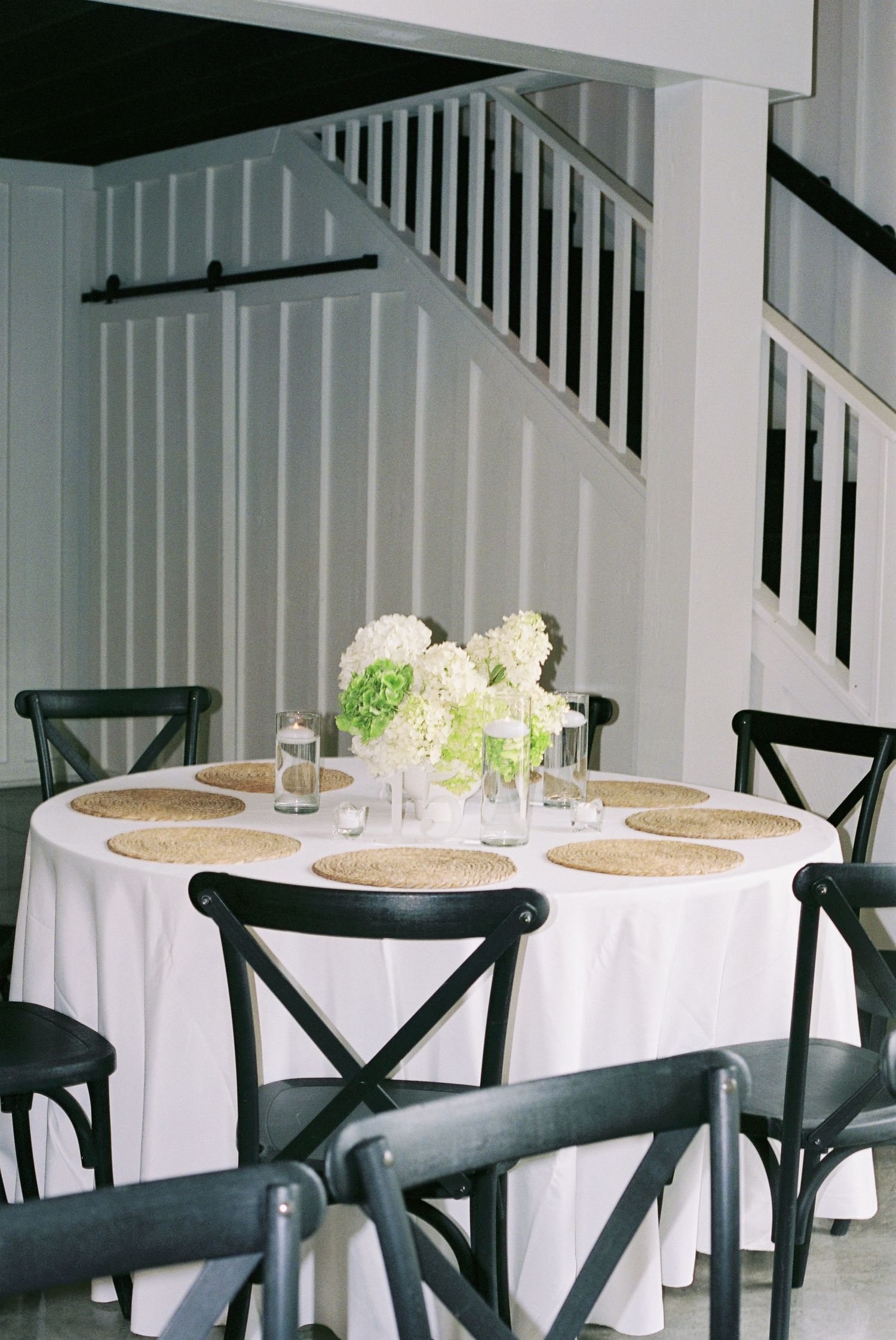 Elegant round dining table with white linen, wicker placemats, black cross-back chairs, and white hydrangea centerpiece near staircase.