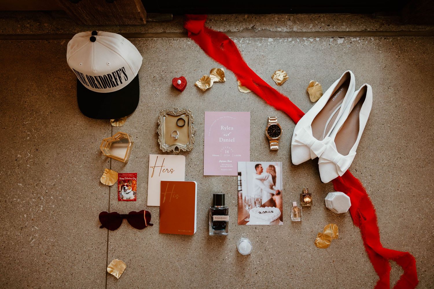Flat lay of wedding details including white bow heels, red ribbon, invitations, sunglasses, hat, jewelry, and perfume on stone floor.