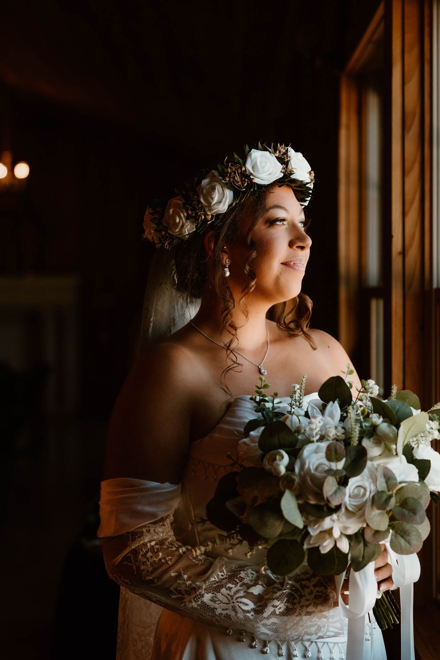 A bride in a white floral crown and strapless gown holds a bouquet of white flowers and eucalyptus by a window.