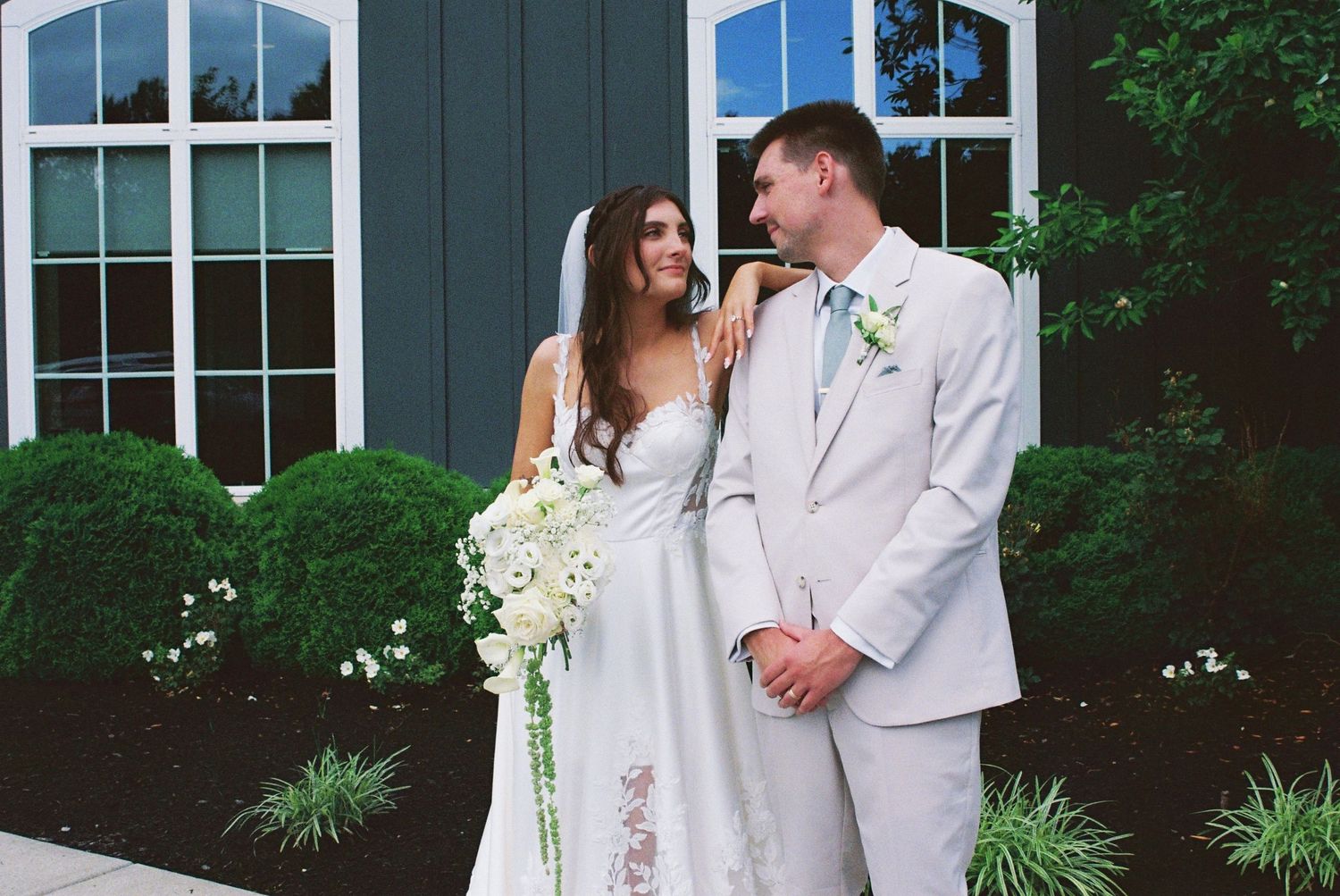 Bride and groom sharing a romantic moment outdoors, bride holding white bouquet, groom in light gray suit.