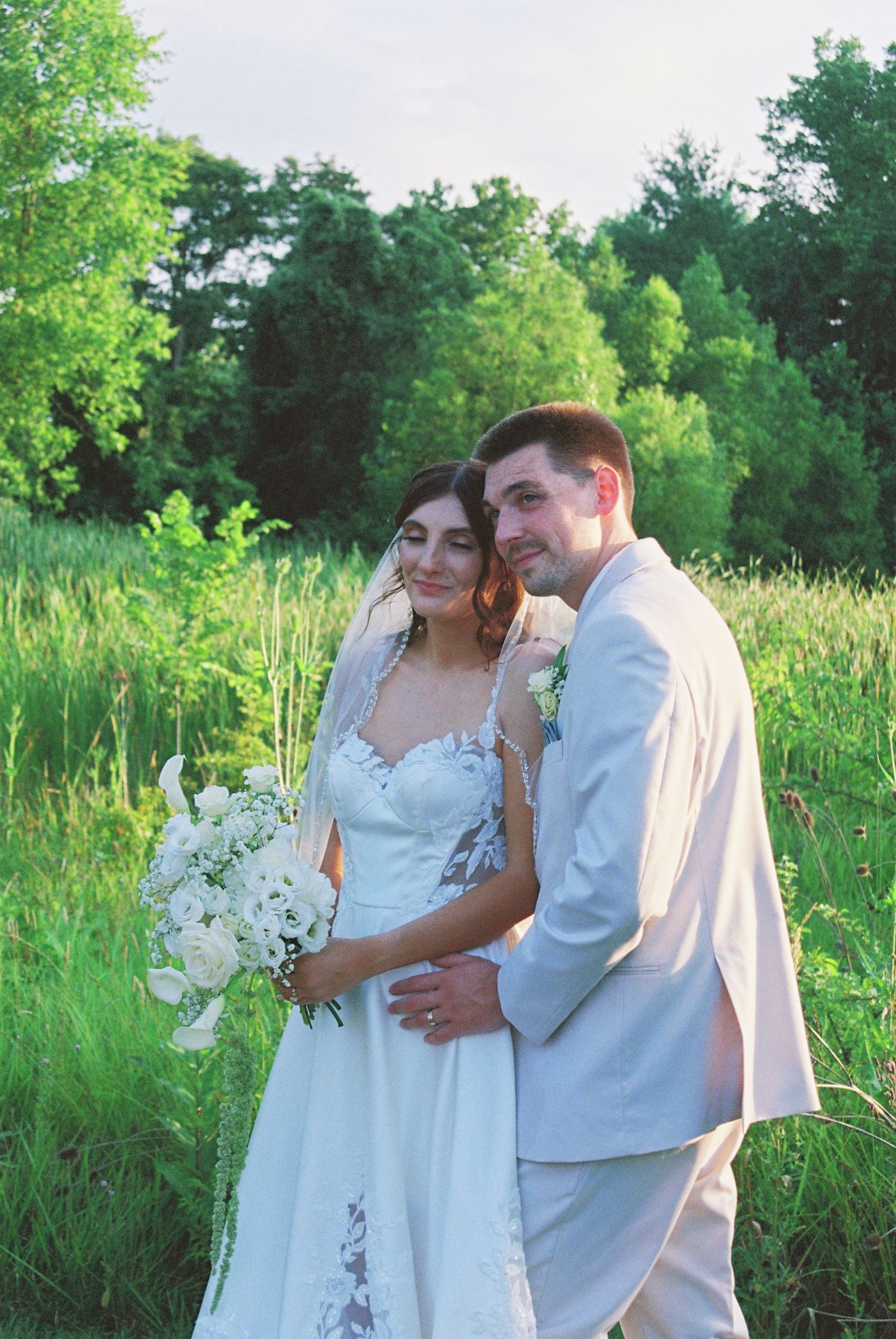 Bride and groom posing together in a sunlit meadow, bride holding a white bouquet.