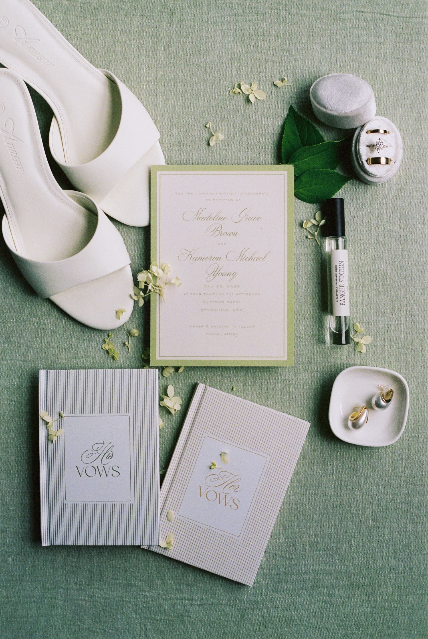 Elegant wedding flat lay with white heels, vow books, invitation suite, rings, and perfume on a sage green background.