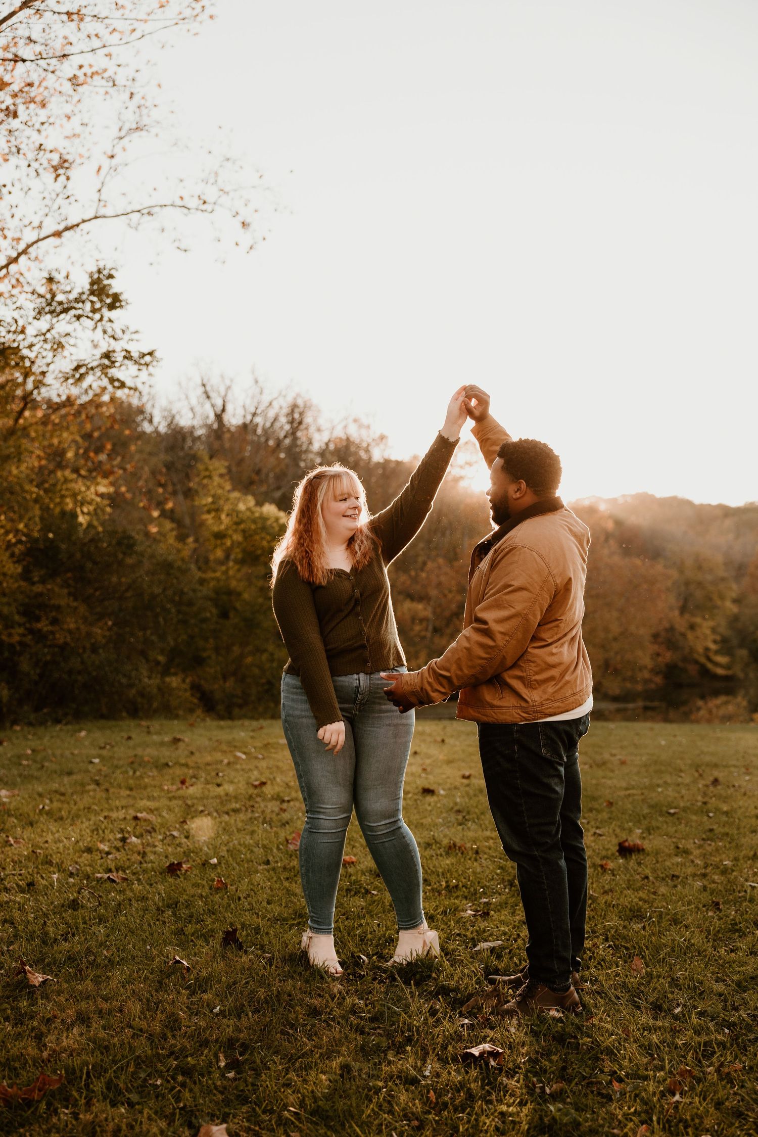 A couple dances playfully in a field during golden hour with autumn foliage in the background.