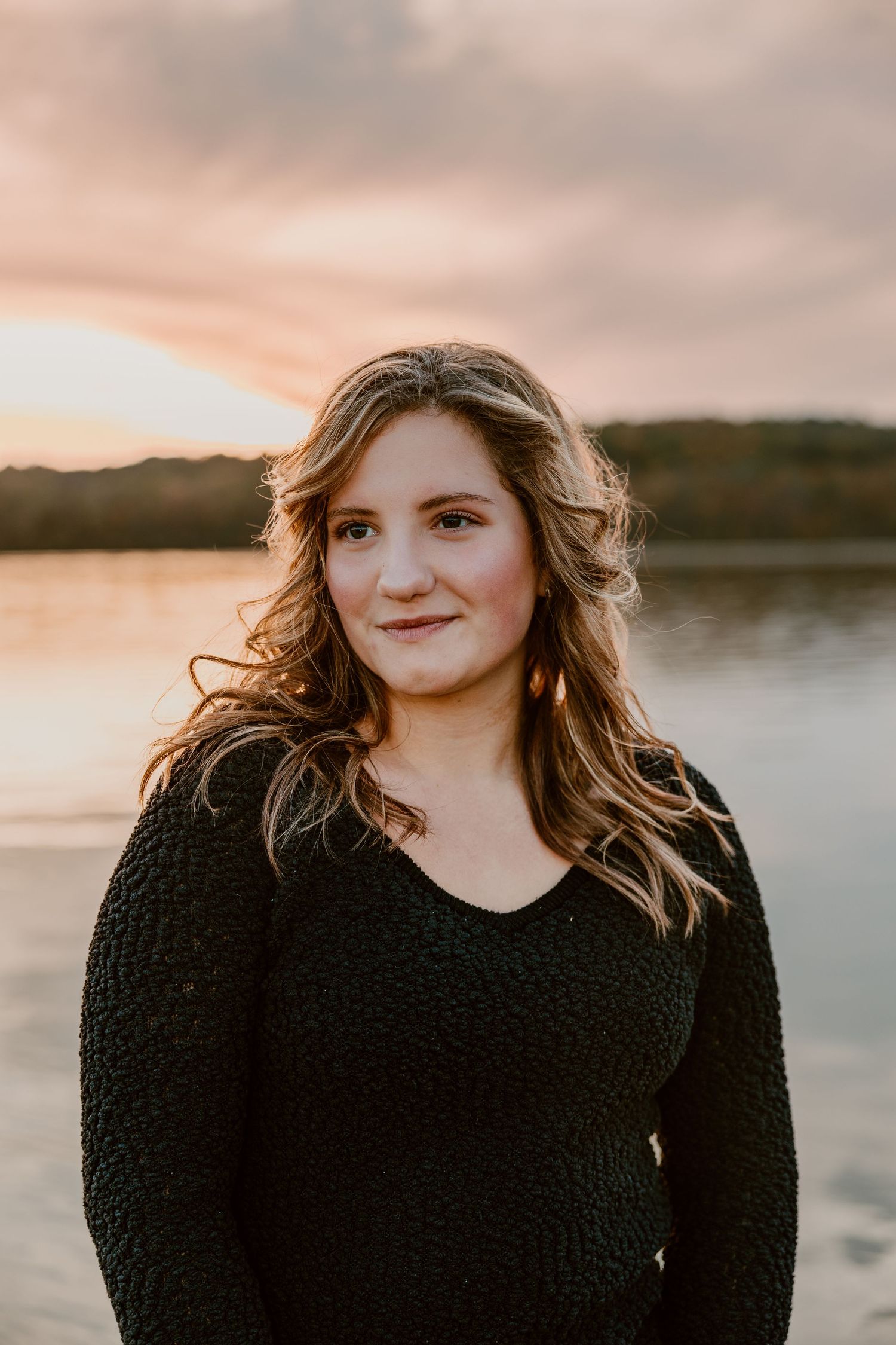 Woman in black sweater poses against sunset lake backdrop with warm lighting.