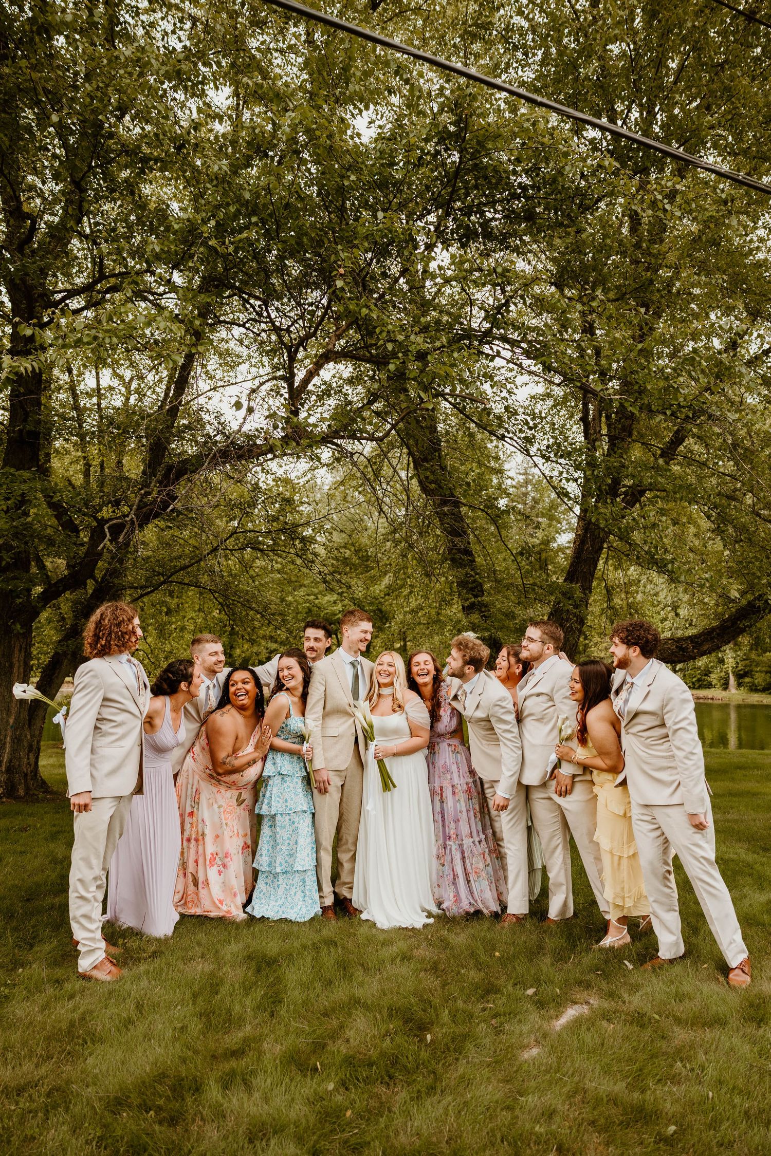 Wedding party in pastel dresses and light suits poses together on a grassy hillside.