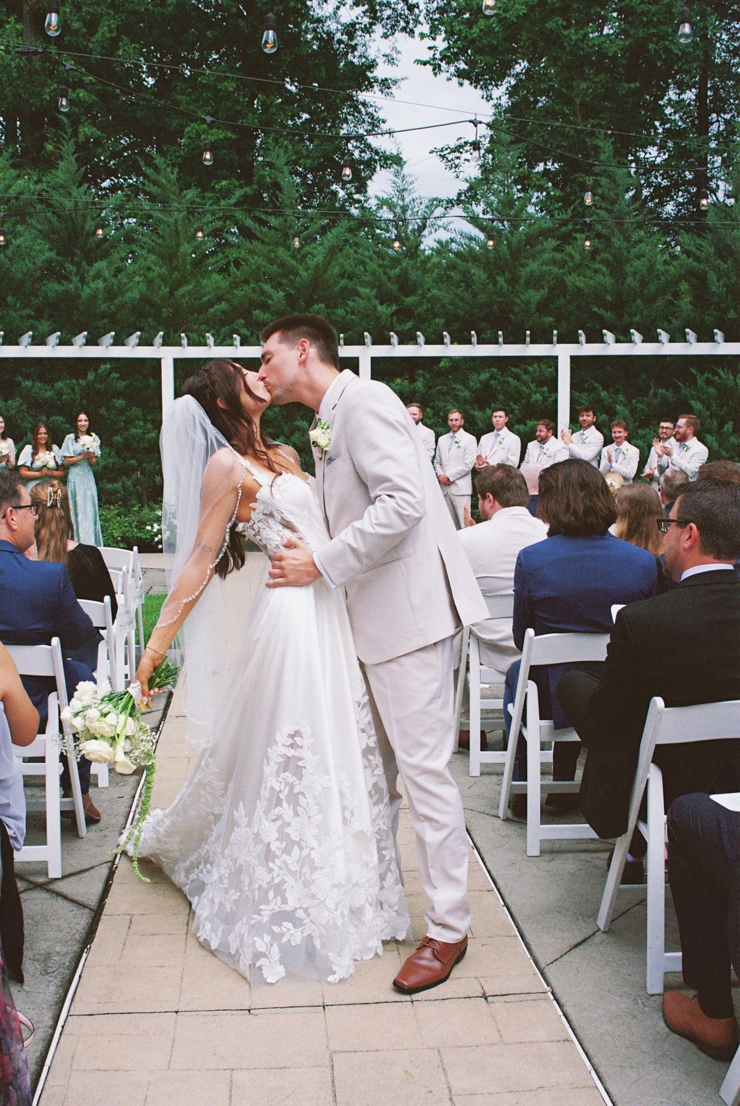 Bride and groom sharing first kiss at outdoor wedding ceremony with seated guests and white chairs.