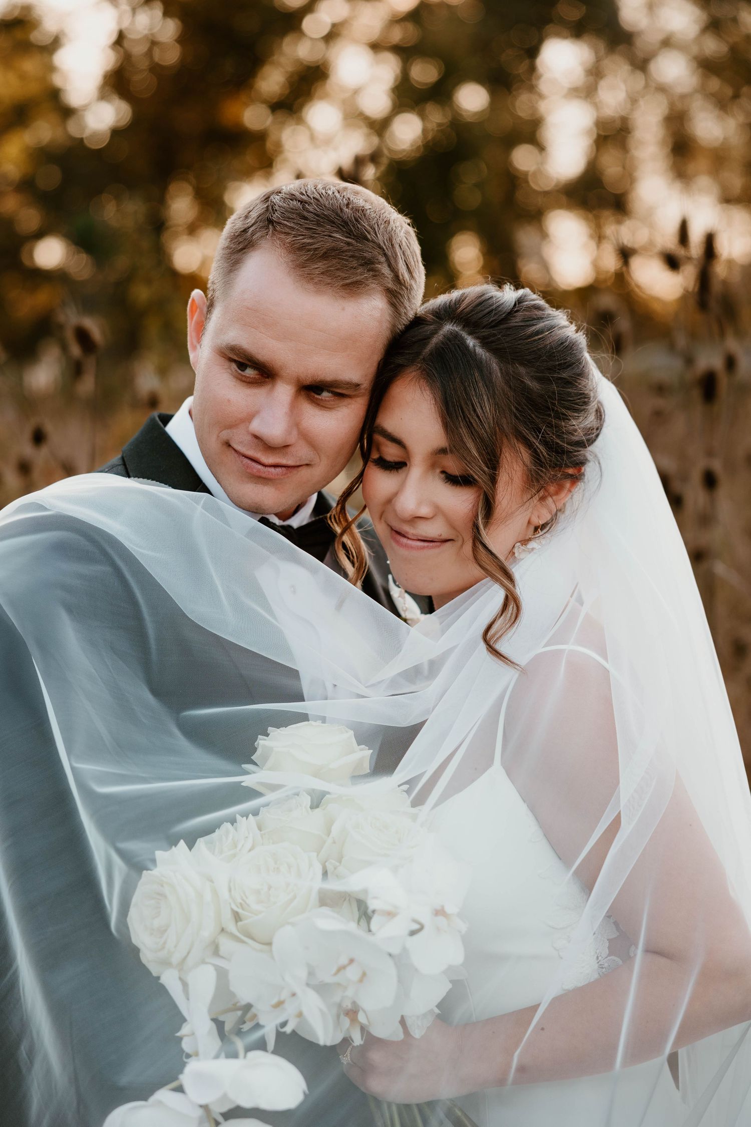 Intimate moment between newlyweds wrapped in flowing white veil during golden hour.