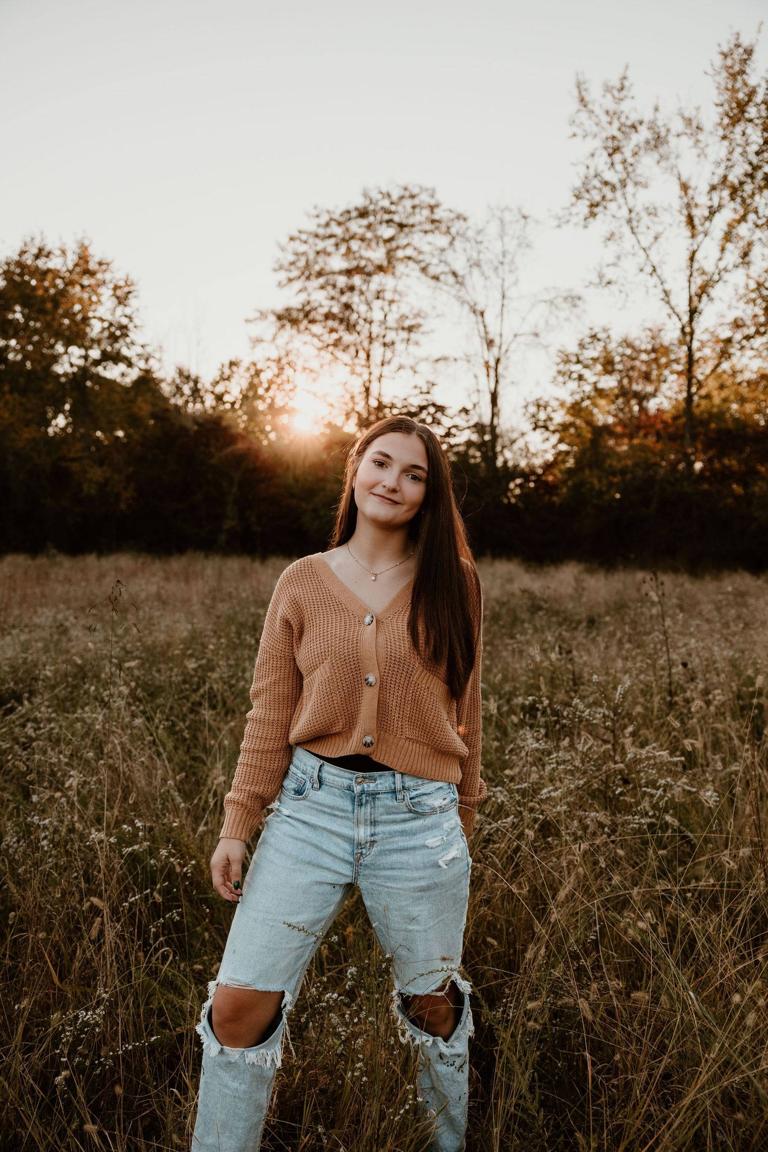 Person in brown cardigan and distressed jeans poses in golden hour field setting.