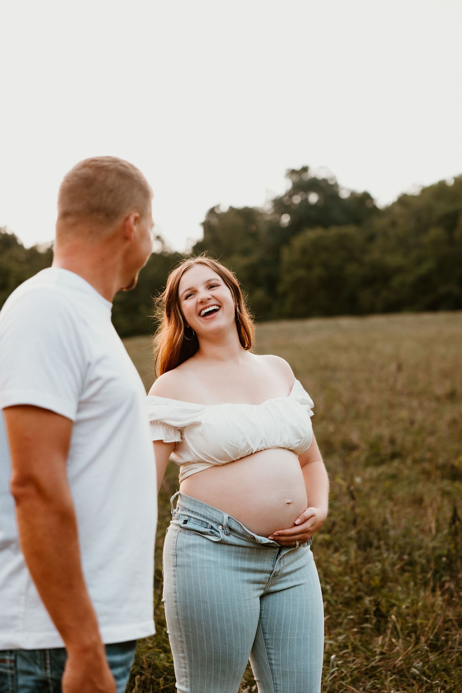 A couple shares a joyful moment in a sunlit field during a maternity photoshoot.