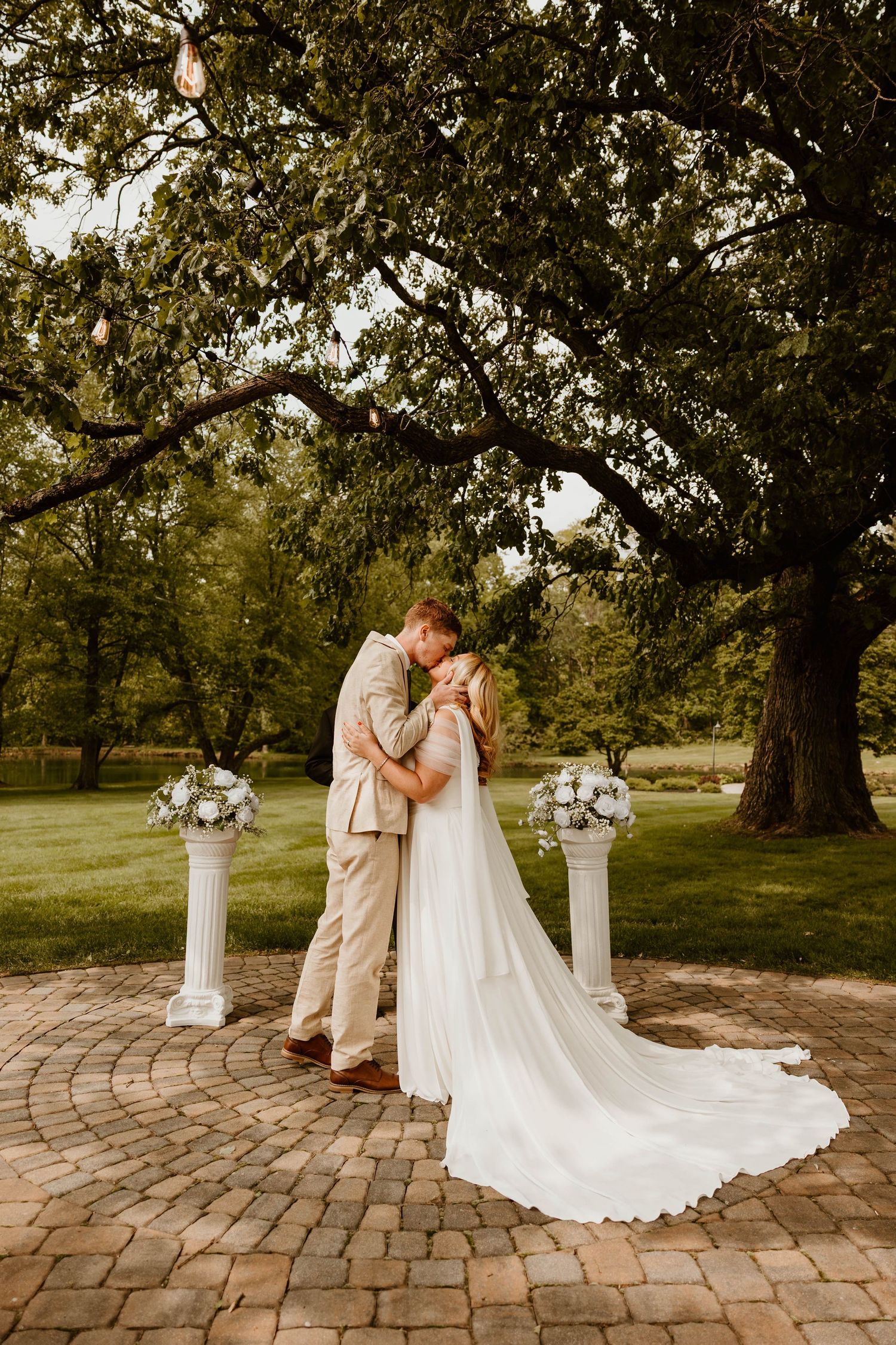 Wedding couple embracing on circular brick patio under majestic oak trees.