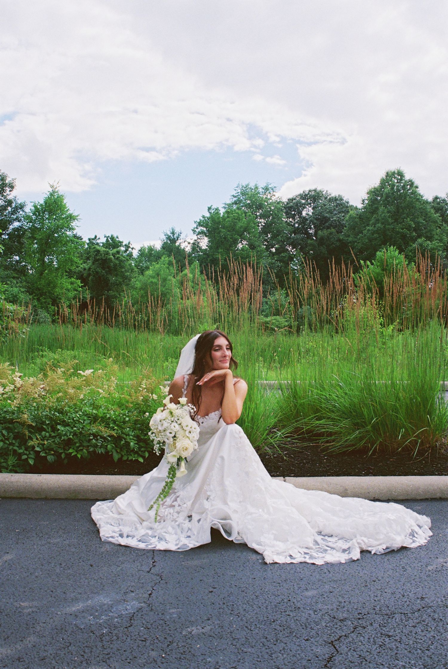 Bride in white lace gown and veil sits on curb holding bouquet with lush green landscape background.