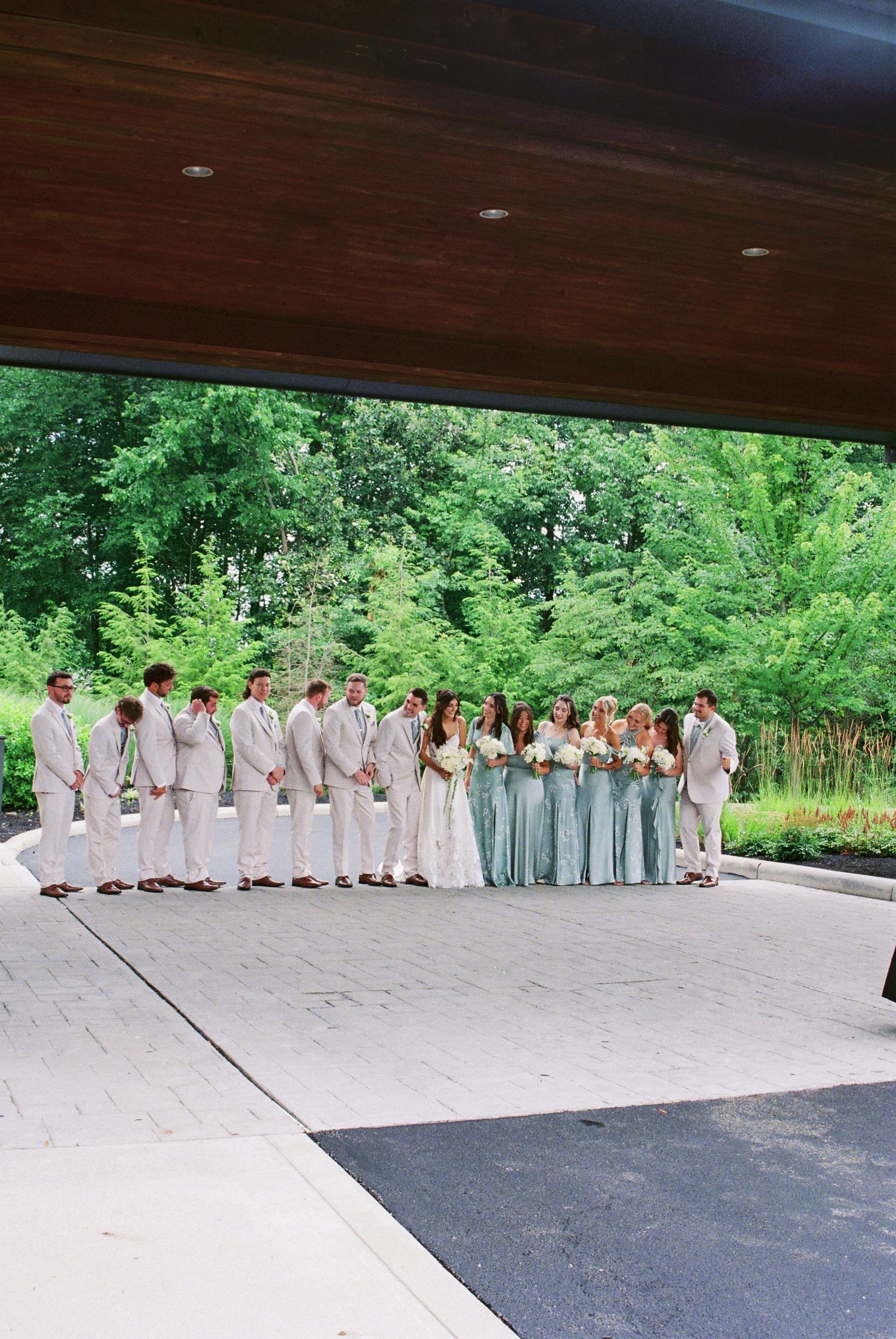 Wedding party in sage bridesmaid dresses and tan suits lined up outdoors under a pavilion with lush green trees.