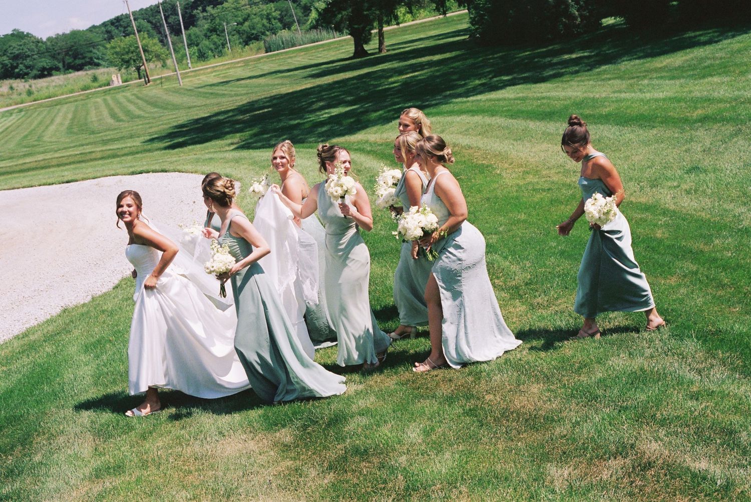 Six bridesmaids in white dresses holding bouquets pose playfully on a green lawn at an outdoor wedding celebration.