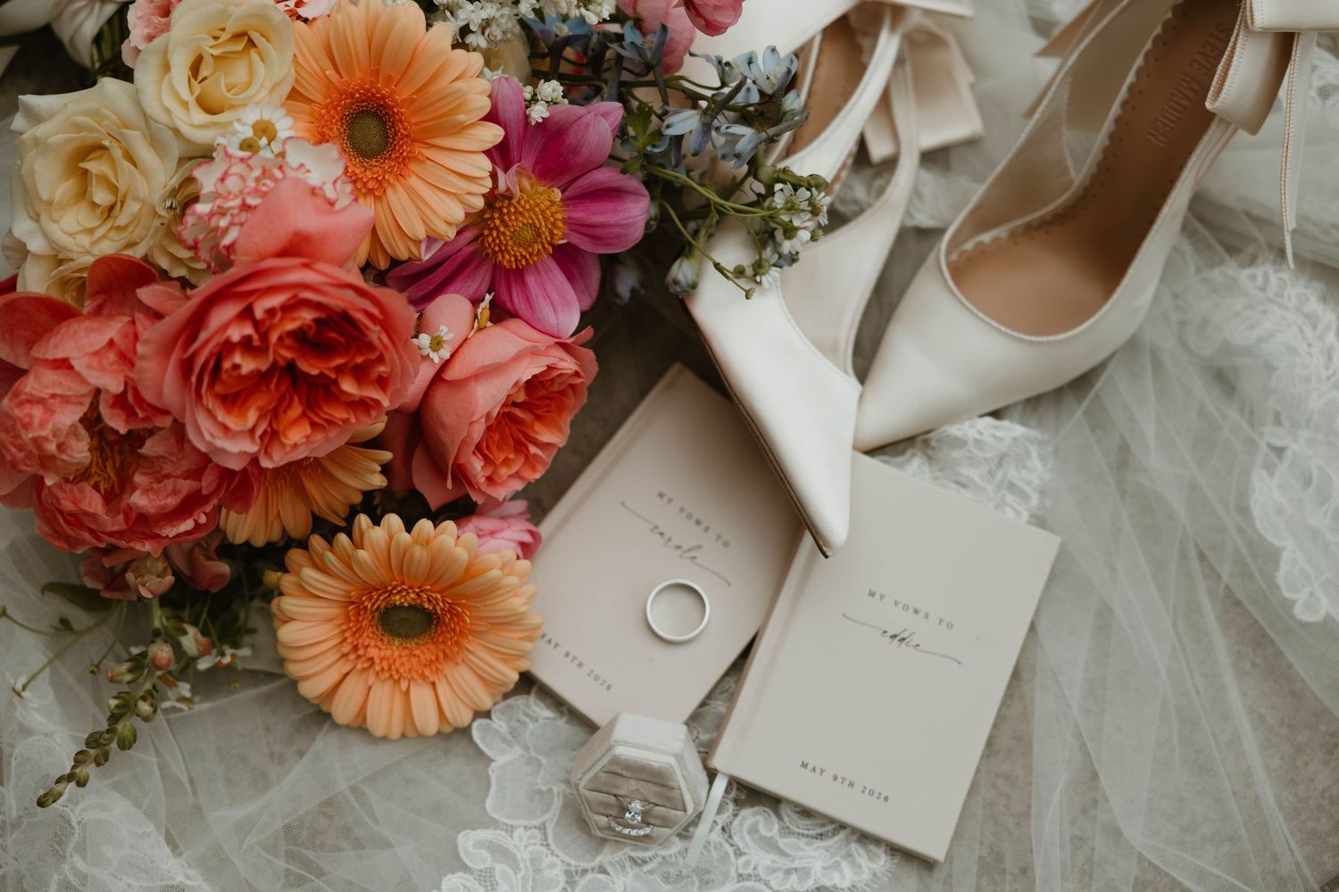 Bridal flat lay with colorful floral bouquet, white heels, wedding rings, and stationery on lace fabric.