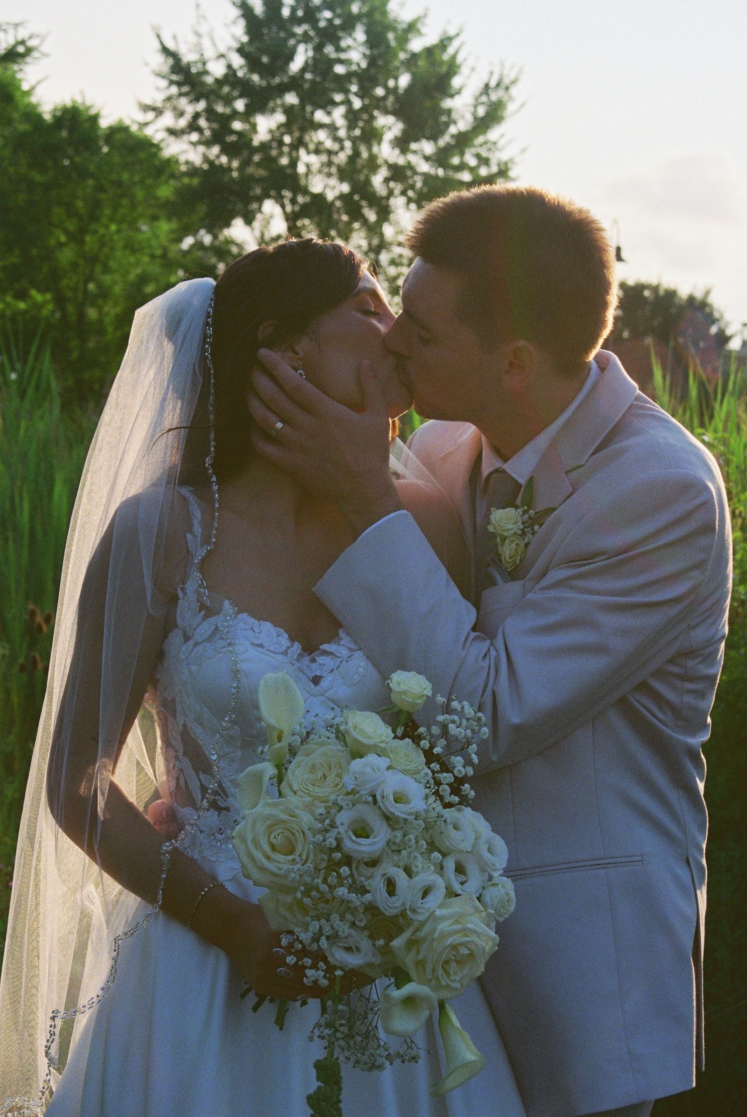 Bride and groom sharing a romantic kiss outdoors at sunset, bride holding white rose bouquet wearing veil.