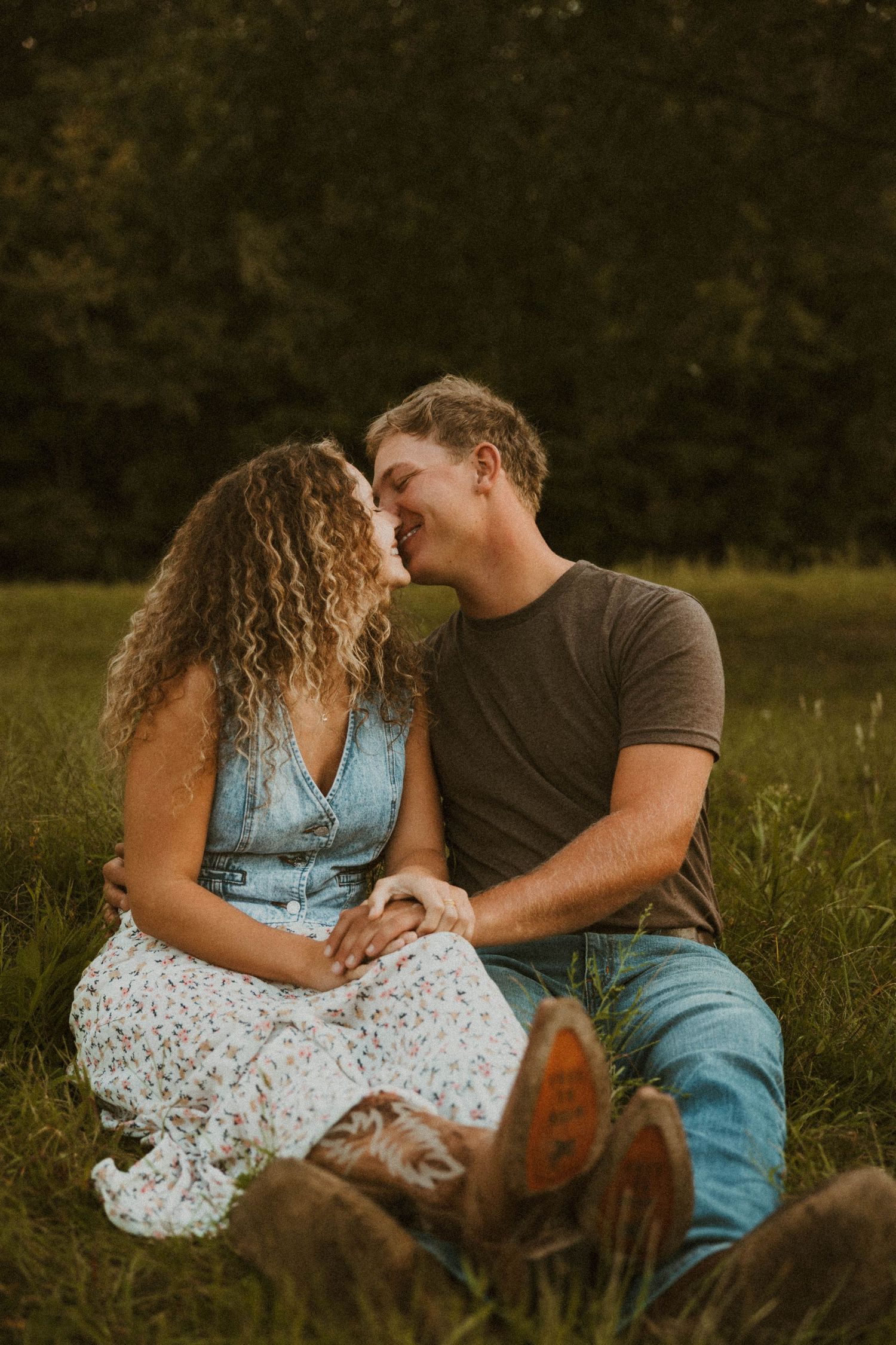 A couple sits together in a grassy field at sunset sharing an intimate moment while their dog rests nearby.