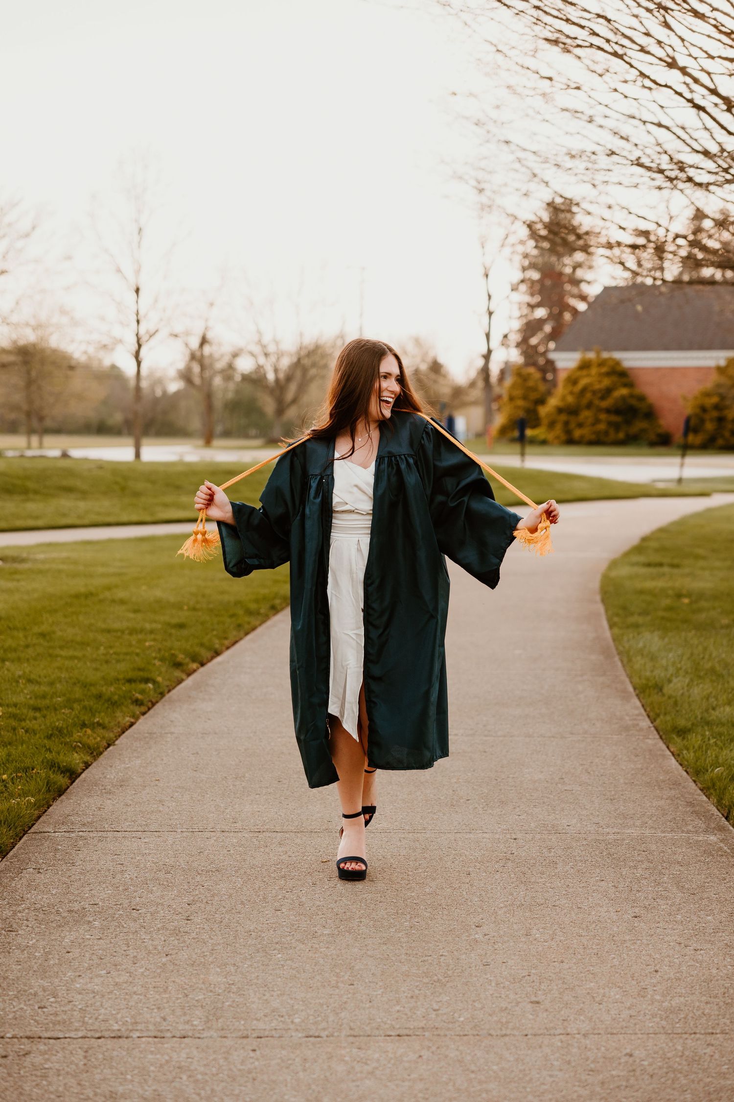 Graduate in black gown twirls on campus sidewalk during sunset.